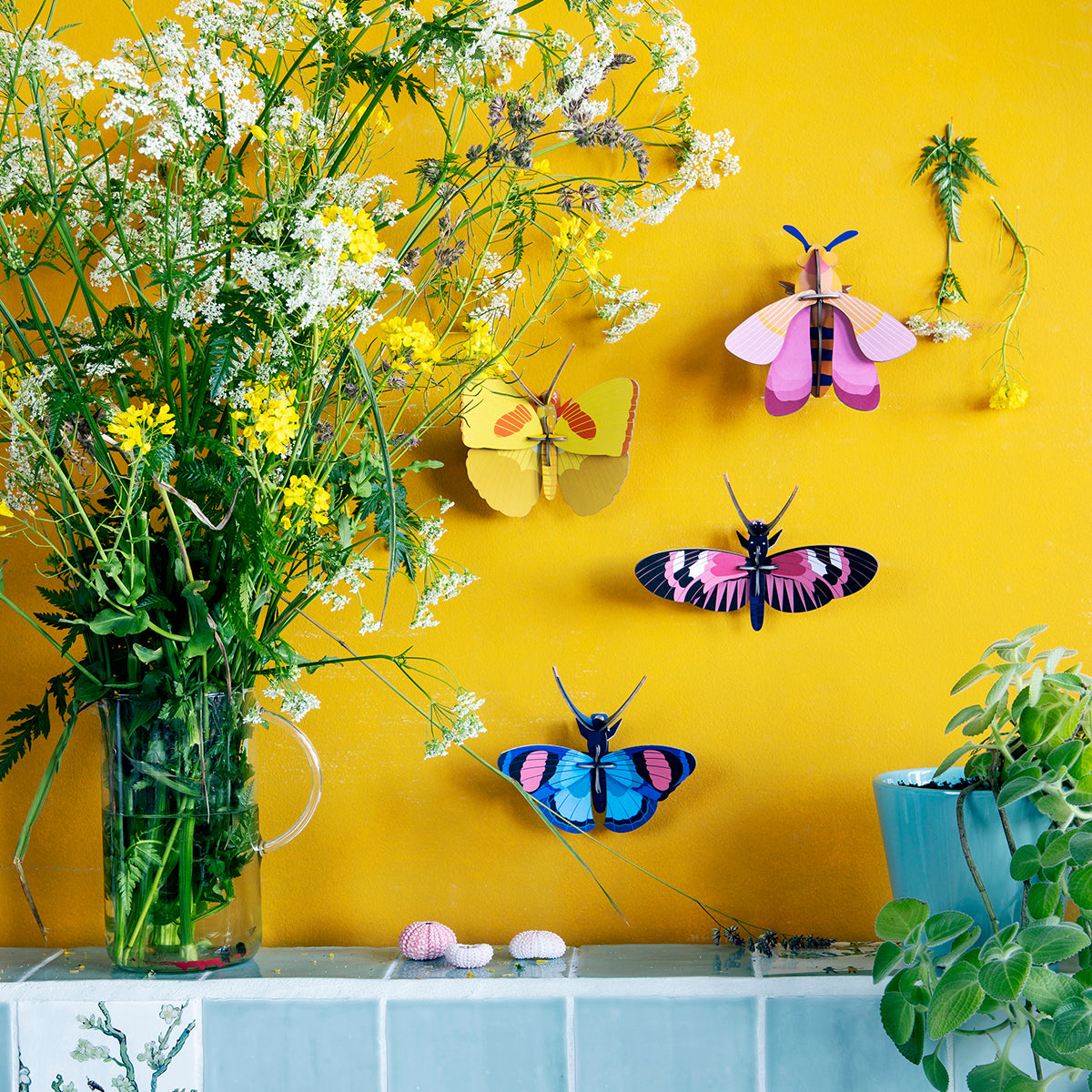 Studio Roof B7 yellow butterfly wall art and other butterflies on bright yellow wall next to a tall vase with white green yellow flowers
