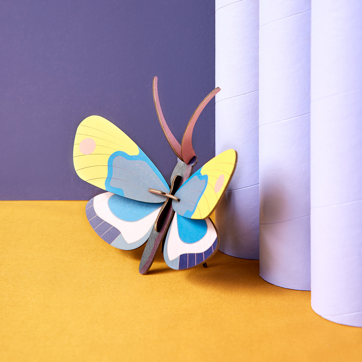 Close up of Studio Roof B7 yellow monarch butterfly wall art on bright yellow surface with deep purple wall and leaning on pale purple pole 