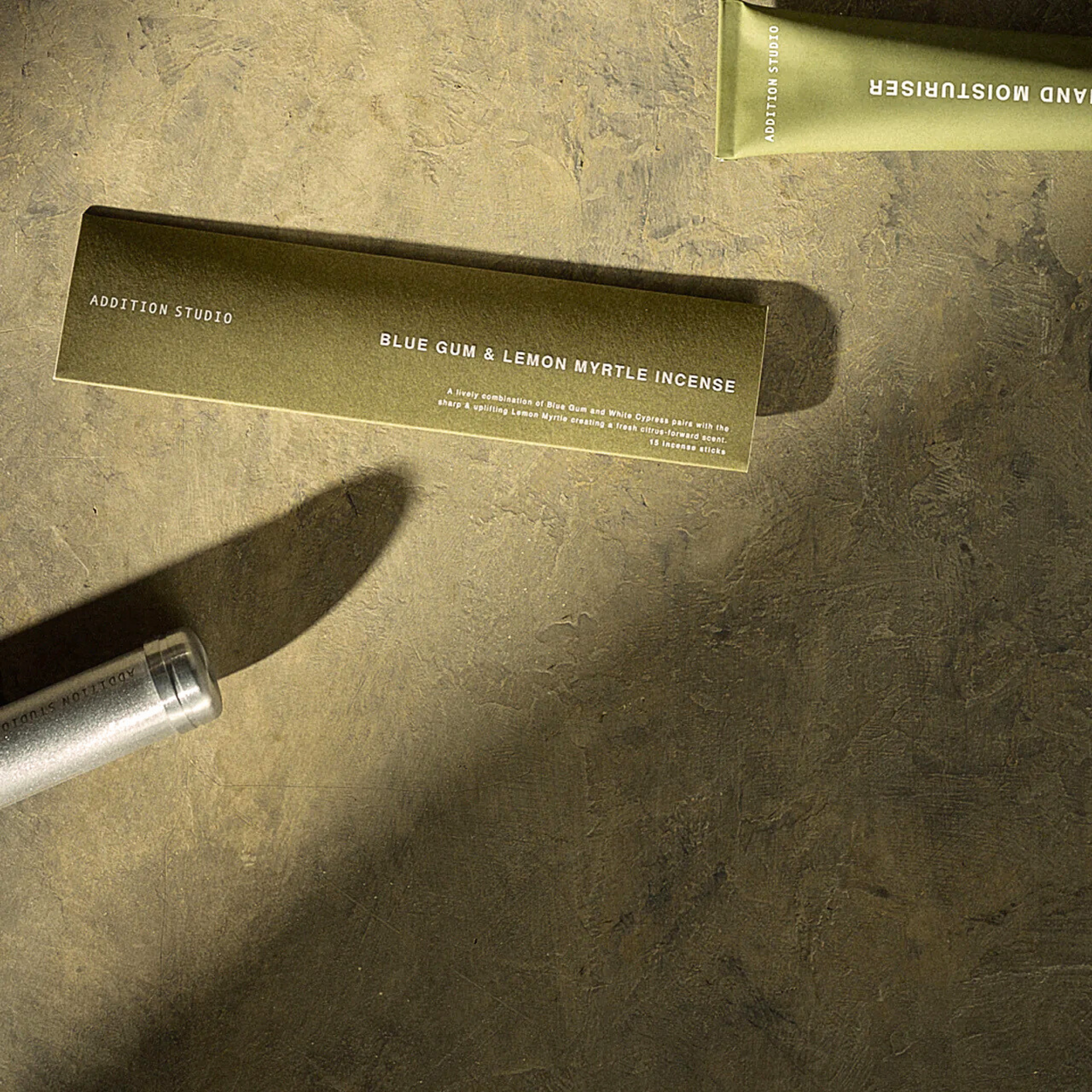 On a surface with a moisturizer, the Addition Studio Incense Refill Blue Gum & Lemon Myrtle, a 15 stick incense in a green box with fresh blend of rosalina, may chang, atlas cedarwood, and white cypress.