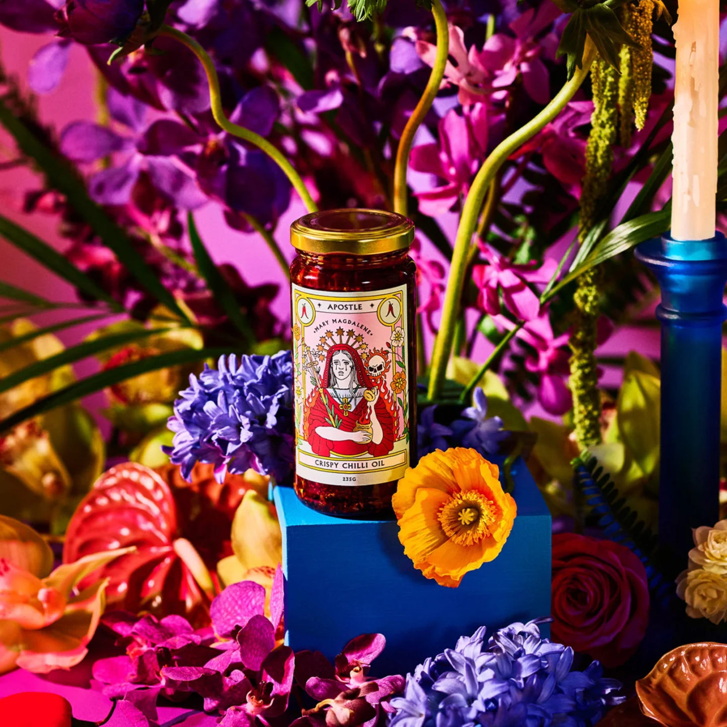 On a blue cube surface with flowers around it, the bottle Apostle Mary Magdalene Crispy Chilli Oil 250ml inspired by flavours across the globe, a mouth-watering blend of crisp, fragranr spices and deep savoury flavour.