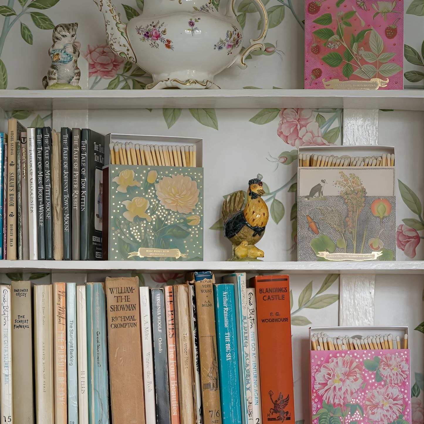 Seen with books and figurines on a shelf, the Archivist Luxury Matches Chien Botanical Drawing which features a green and orange floral illustration with a collie dog and gold foil detail by Florence Balducci. Contains 125 matches (100 mm, white tips). Litho printed in the UK with FSC-certified wood and wrapped in compostable cello-ideal for coffee tables, mantelpieces, or gifting.