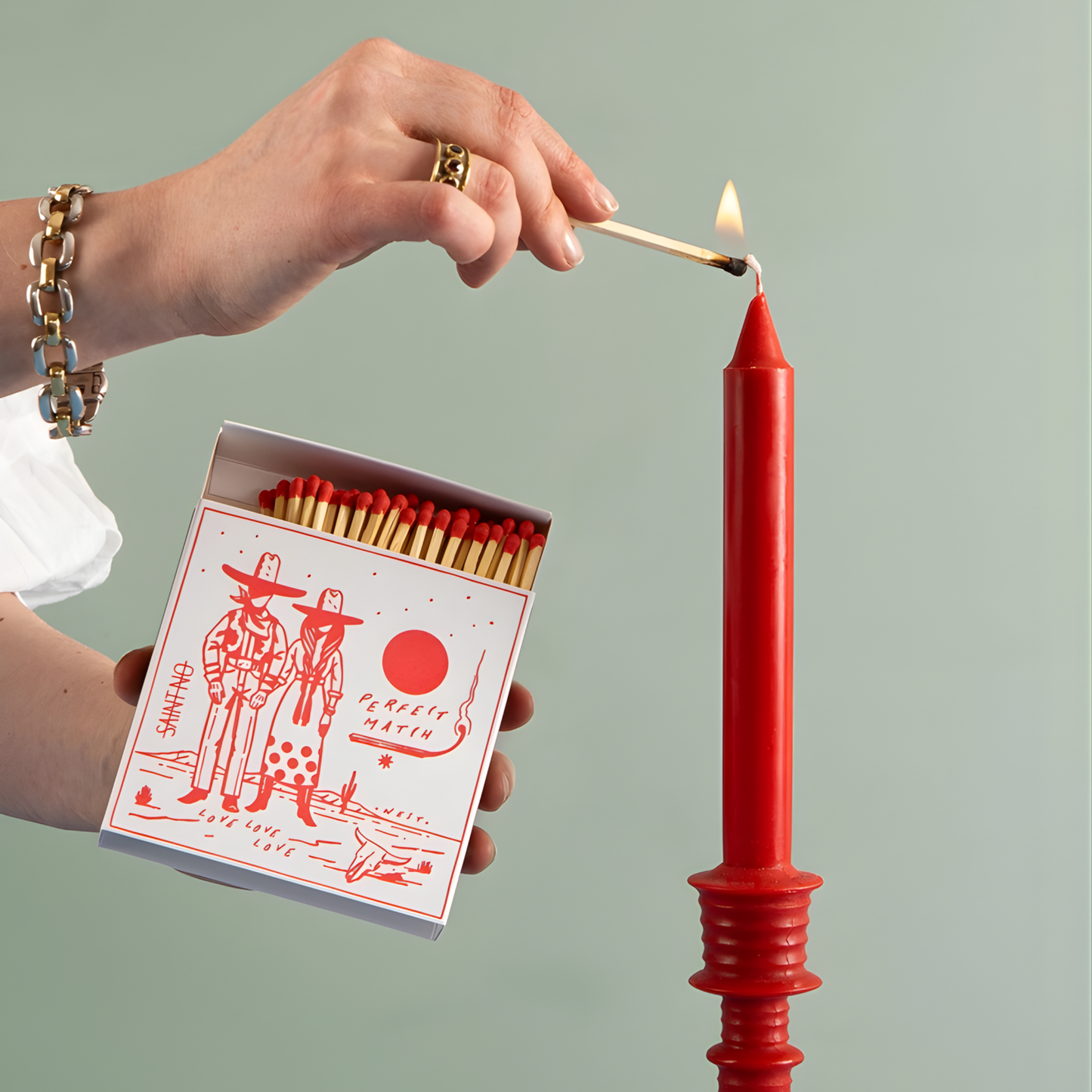 Lighting a red candle with the Archivist Luxury Matches Cowboy Perfect Match: Features a Western scene of a cowboy and cowgirl beneath the sun with the words "Perfect Match, Love love, love." Contains 125 matches (100 mm, red tips), letterpress printed in the UK with FSC-certified wood and wrapped in compostable cello-perfect for coffee tables, mantelpieces, or gifting.