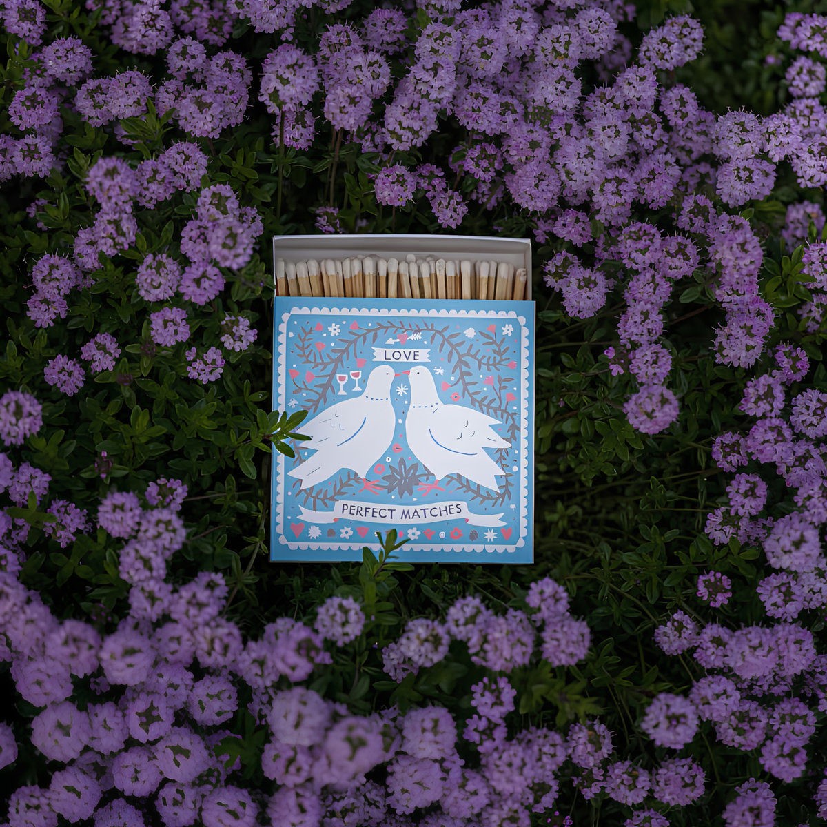 Archivist Luxury Matches Dove Perfect in blue and white, FSC Mix certified paper matchbox with two white doves, 125 natural wooden matches with yellow tips, shown on top of the purple flowers.