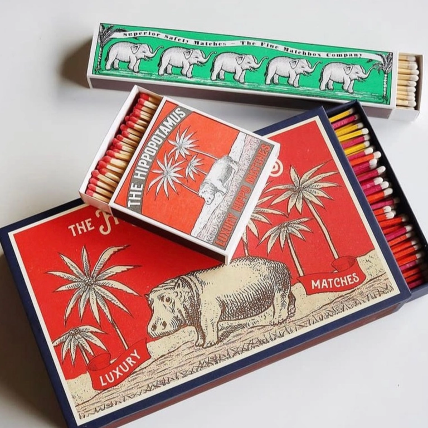 On a big match box, open and showing the red tipped matches from Archivist Hippopotamus Matches - Square Matchbox with Luxury Hippo and Palm Tree Design - 125 Natural Red-Tipped Matches - FSC Wood, UK Letterpress Printed, Eco-Friendly Decorative Match Set