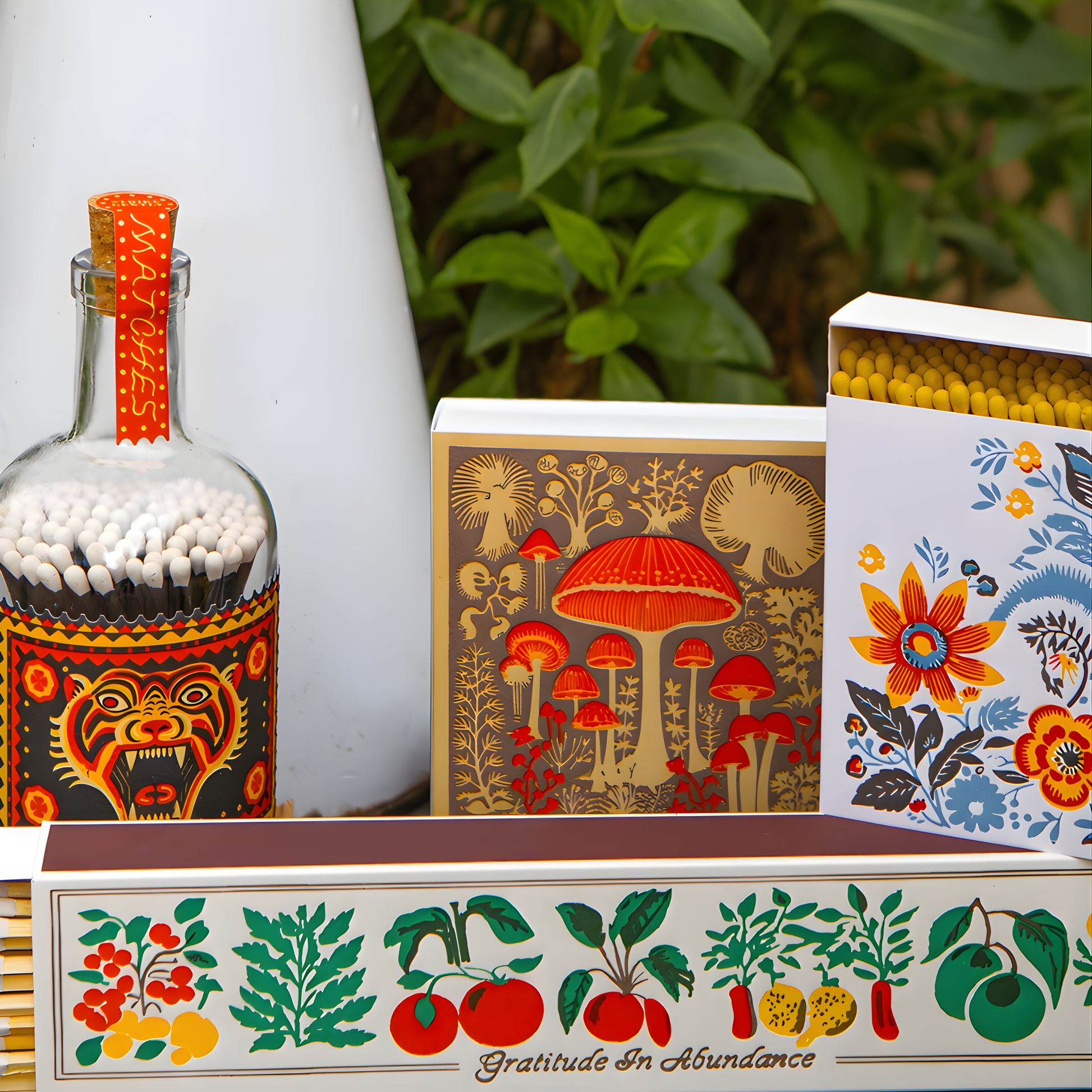 Showing different match containers on a surface with the Archivist Luxury Matches Mushroom which features a selection of wild mushrooms in neutral tones with red accents. Contains 125 matches (100 mm, red tips, white tray), letterpress printed in the UK with FSC-certified wood and wrapped in compostable cello-ideal for coffee tables, mantelpieces, or gifting.