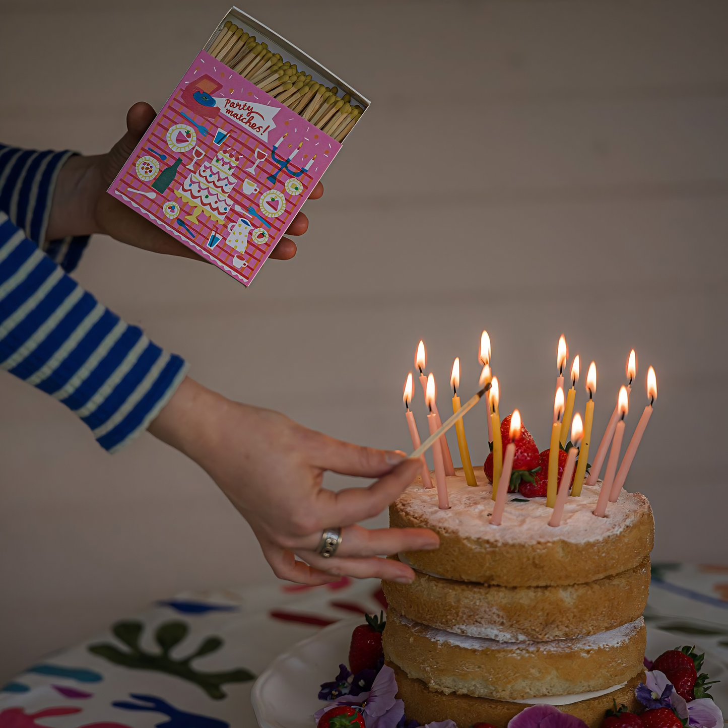 A woman lights the candles on a cake using the Archivist Luxury Matches featuring an illustration Picnic Party with cakes and drinks, 'Party Matches' text.