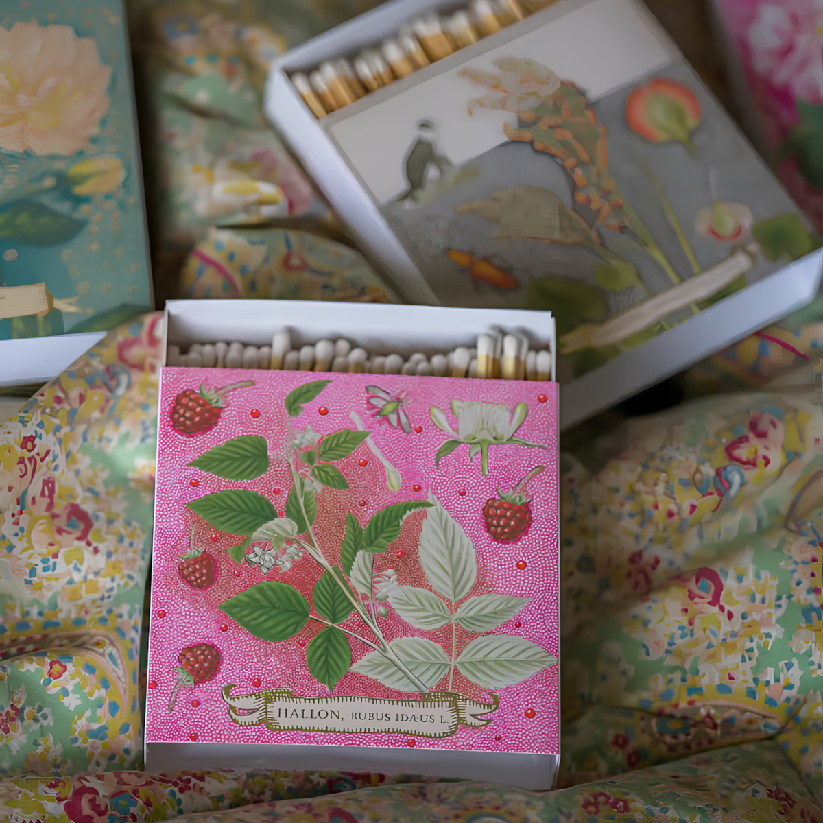 Shown lying with other match designs on a cloth, the Archivist Luxury Matches Rose Framboise in pink and green, FSC Mix certified paper matchbox with raspberry and leaf illustrations, gold foil details and 125 natural wooden matches with white tips.
