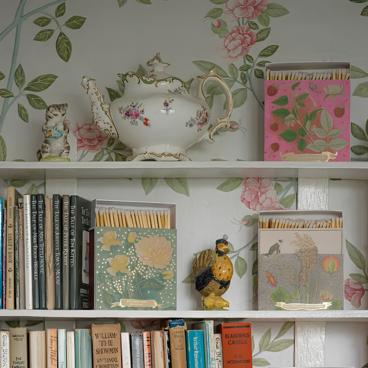 Archivist Luxury Matches Rose Framboise in pink and green, FSC Mix certified paper matchbox with raspberry and leaf illustrations, gold foil details and 125 natural wooden matches with white tips, shown on a shelf with books, other matches, figurine and a kettle.