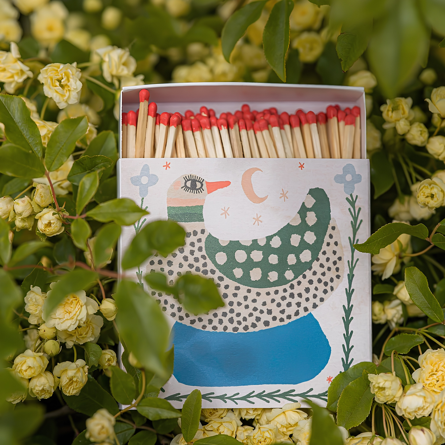 On top of the little yellow flowers, the Archivist Luxury Matches featuring an illustration of a Spotted Duck with green wings sitting in a pond beneath the moon.