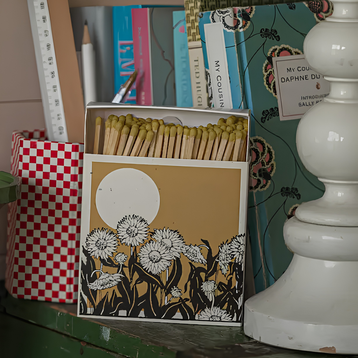 Close up of Archivist Luxury Matches Sunflower Fields in yellow and beige, FSC Mix certified paper matchbox with sunflower field illustration, containing 125 natural wooden matches with yellow tips, shown on top of a wooden cabinet with a lamp beside it.