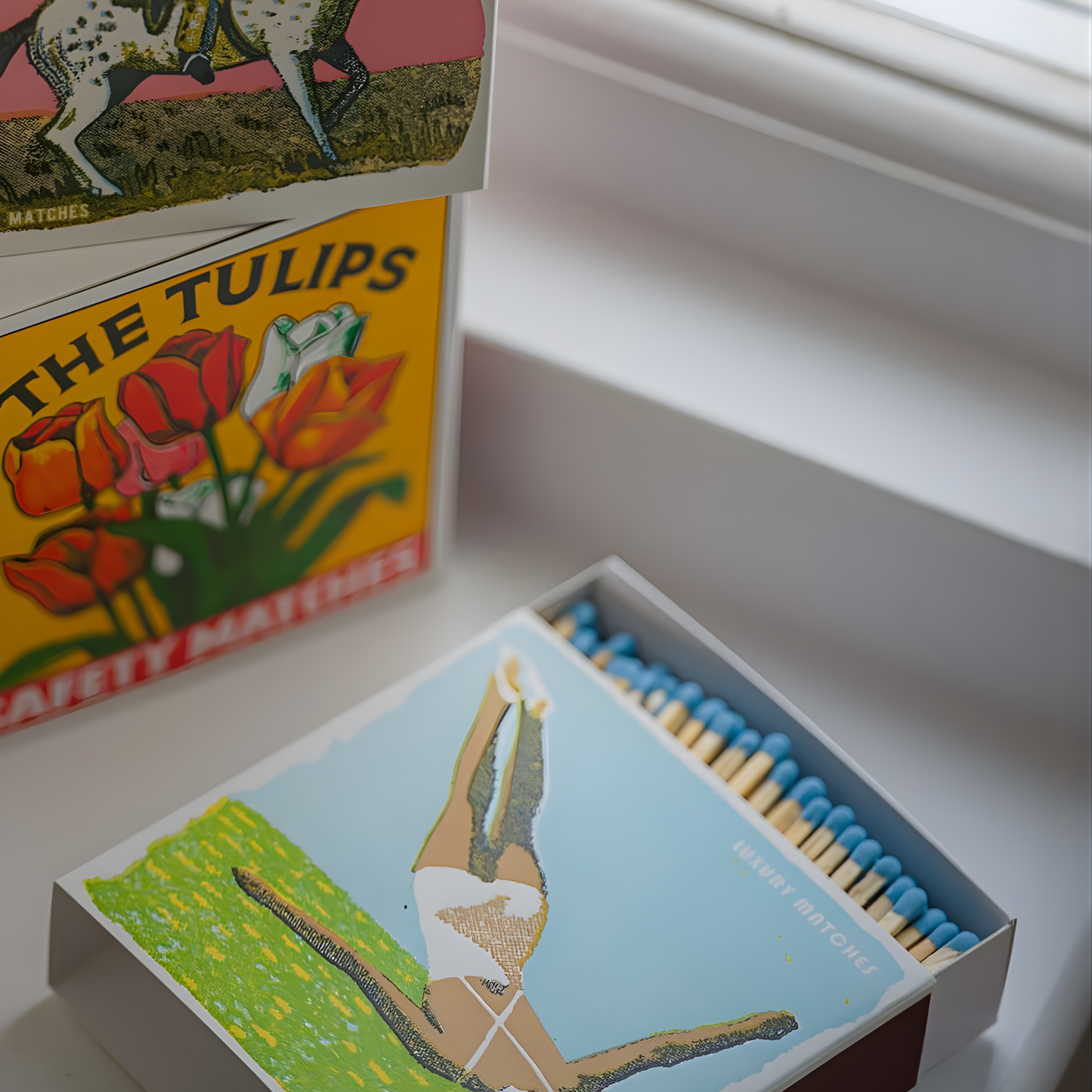 Seen near the window ledge, the Archivist Luxury Matches The Diver which features a vintage-inspired illustration of a woman diving elegantly into the water. Contains 125 matches (100 mm, blue tips, white tray), letterpress printed in the UK with FSC-certified wood and wrapped in compostable cello-perfect for coffee tables, mantelpieces, or gifting.