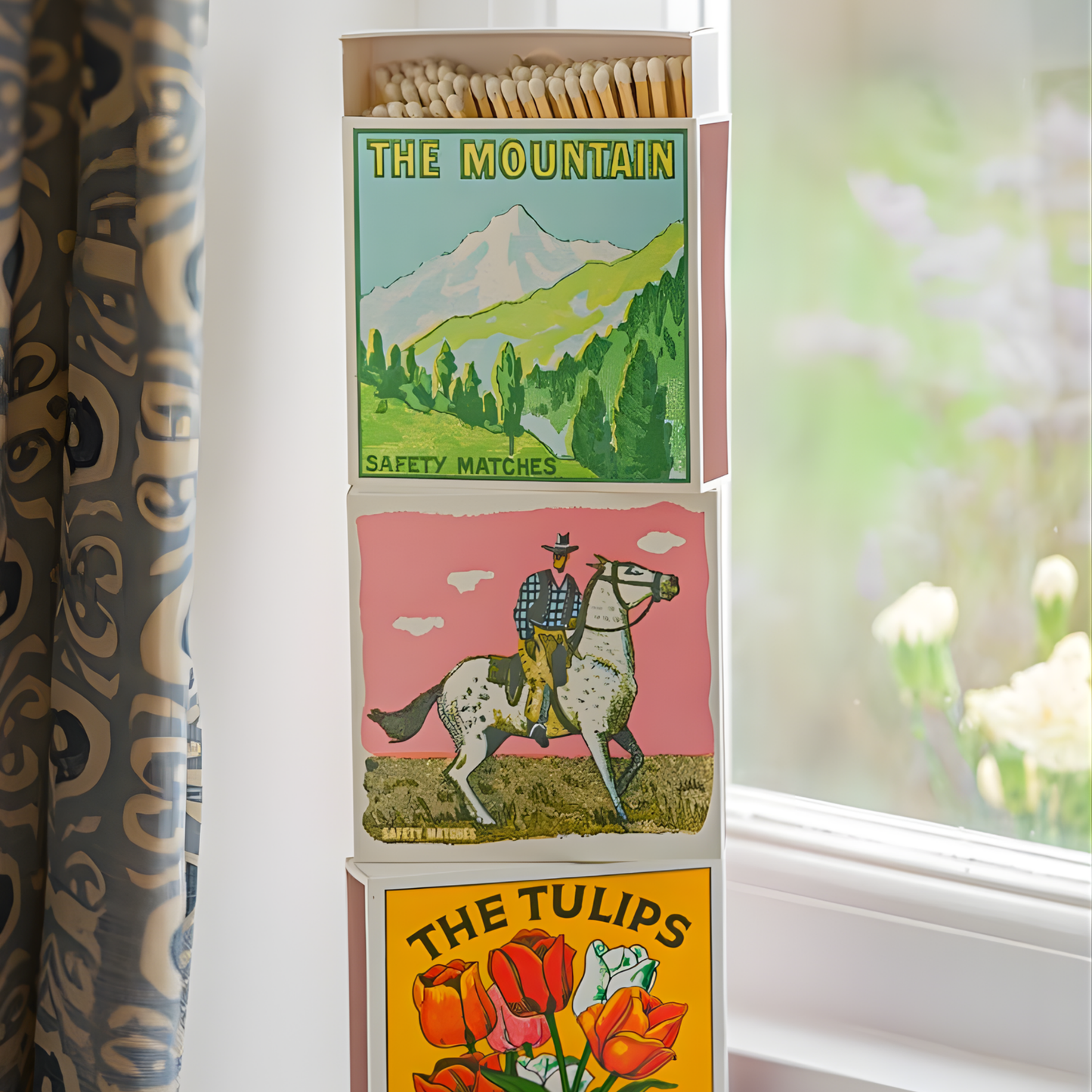 Match boxes being stacked beside the window ledge that includes the Archivist Luxury Matches The Mountain which features a snowy mountain range against a bright blue sky with the words "Safety Matches." Contains 125 matches (100 mm, yellow tips, white tray), letterpress printed in the UK with FSC-certified wood and wrapped in compostable cello-ideal for coffee tables, mantelpieces, or gifting.