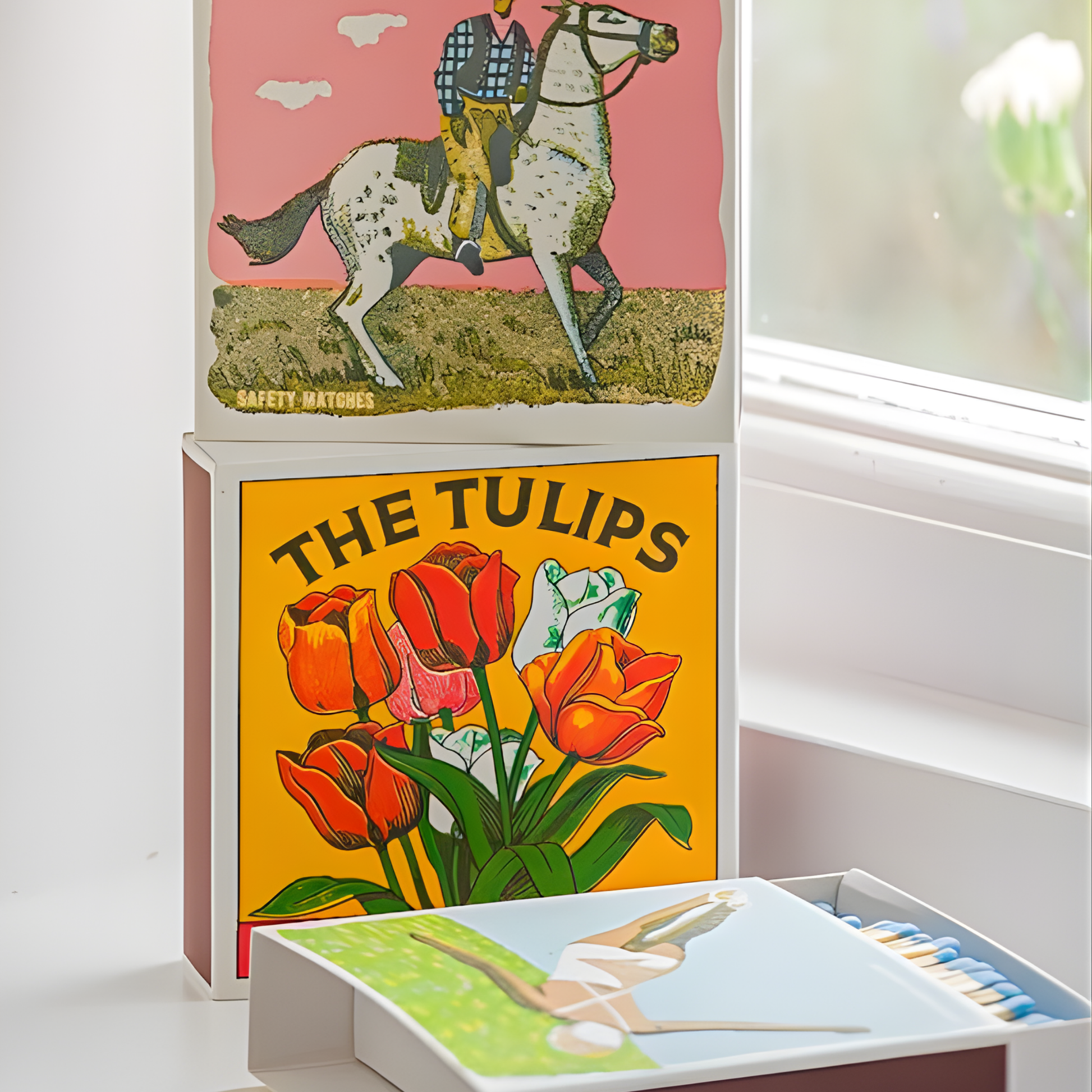 Seen at the bottom of the stacked match boxes, which is the Archivist Luxury Matches The Tulips which features colorful tulips on a bold yellow background with the words "The Tulips, Safety Matches." Contains 125 matches (100 mm, yellow tips, white tray), letterpress printed in the UK with FSC-certified wood and wrapped in compostable cello-perfect for coffee tables, mantelpieces, or gifting.