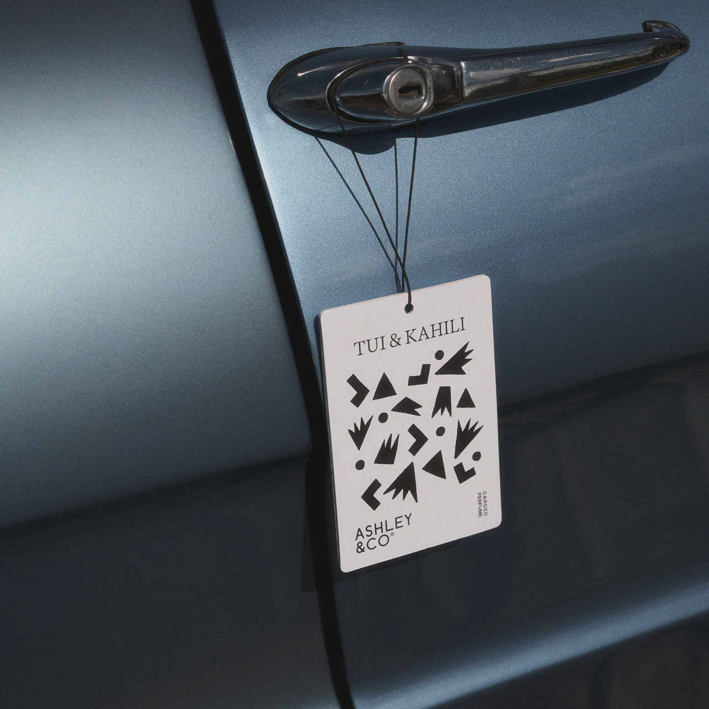 Front view of the Ashley & Co Carded Perfume Air Freshener Tui & Kahili hung on the car door handle.