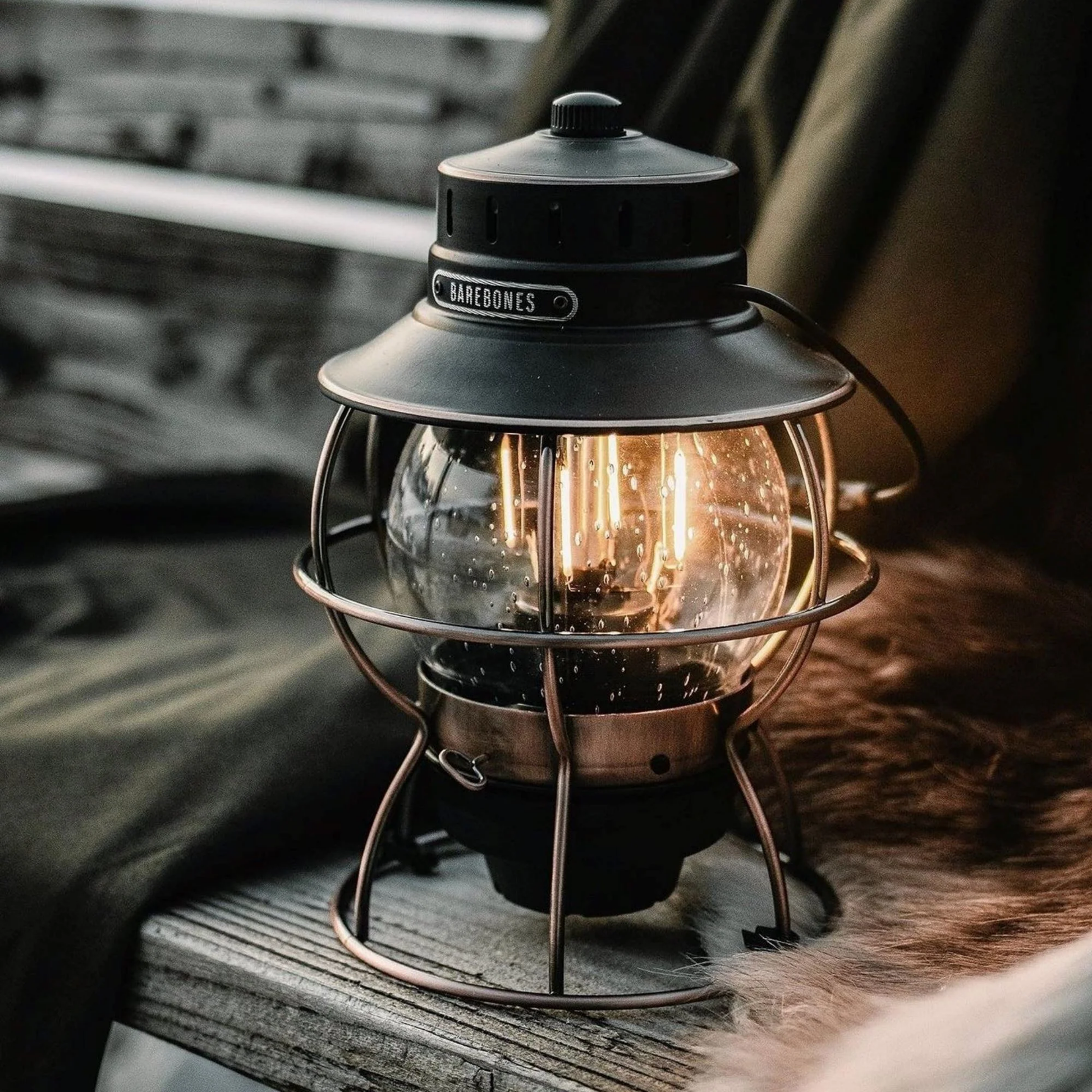 Barebones railway lantern bronze outside on wooden bench next to coats switched on