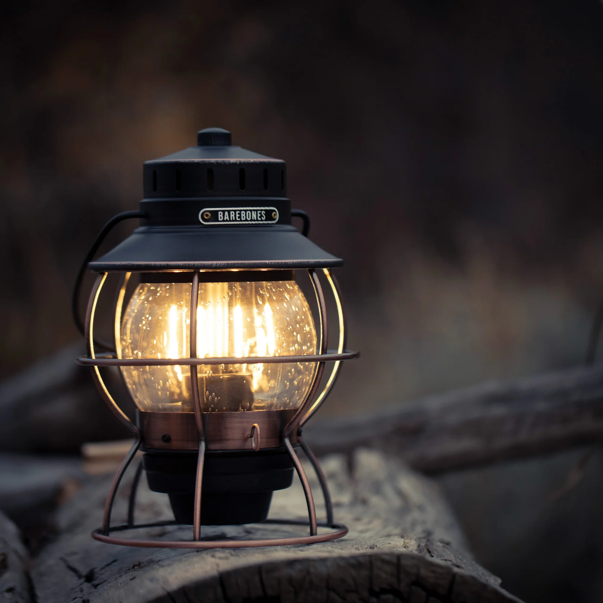 Barebones railway lantern bronze outside on tree log switched on