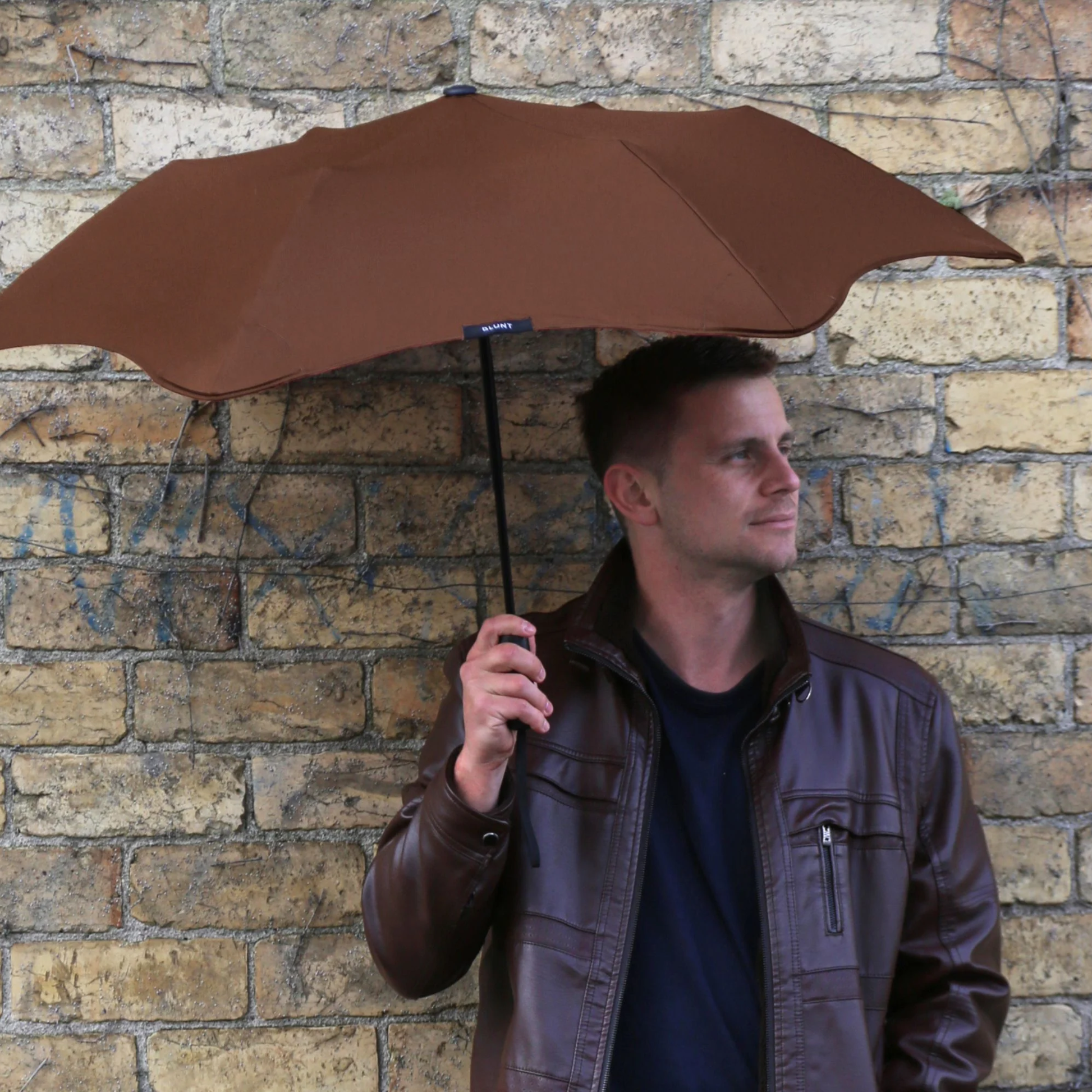 Front view of a man holding the Blunt Umbrella Metro Espresso, that has a Dark and bold smooth edge, a classic tone that adds warmth and depth to rainy days. Compact, collapsible and convenient, consisting of the detachable UV fabric canopy, sturdy frame, shaft and handle. Fits in your handbag, tote or backpack. Built for fast living, those who travel light, and any weather.