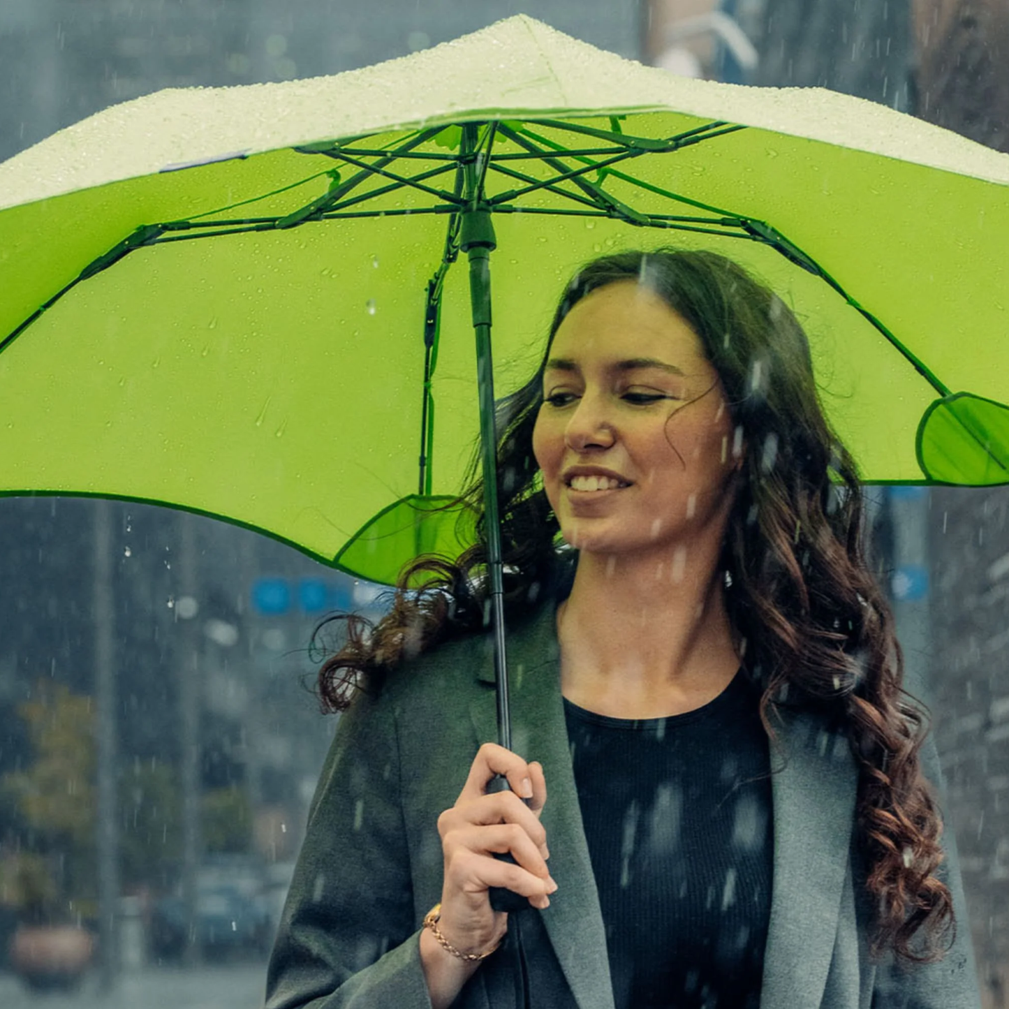 Front view of a woman with the Blunt Umbrella Metro Meadow Green that is compact, collapsible and convenient. Consist of the detachable UV fabric canopy, sturdy frame, shaft and handle. Fits in your handbag, tote or backpack. Built for fast living, those who travel light, and any weather.