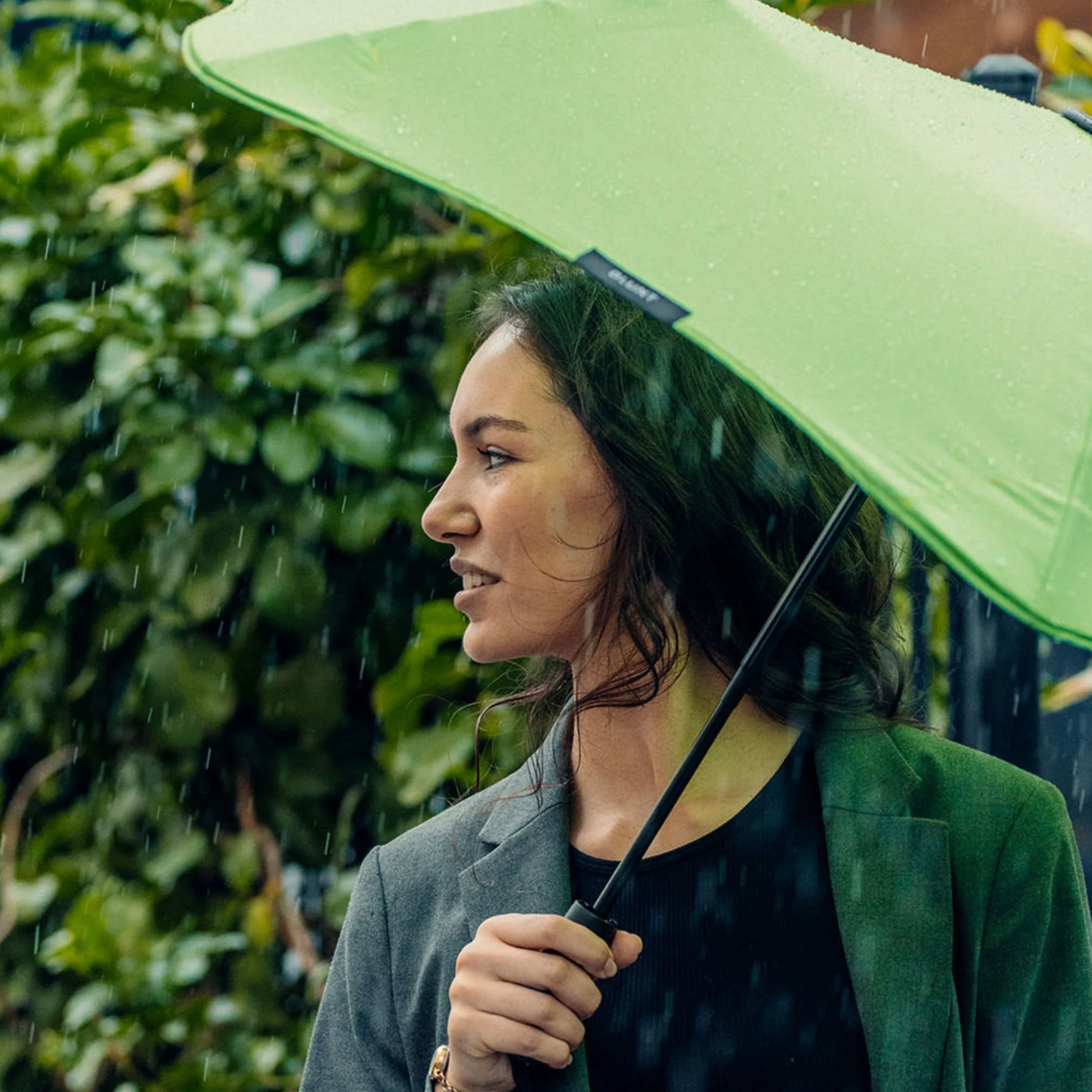 Side view of a woman with the Blunt Umbrella Metro Meadow Green that is compact, collapsible and convenient. Consist of the detachable UV fabric canopy, sturdy frame, shaft and handle. Fits in your handbag, tote or backpack. Built for fast living, those who travel light, and any weather.