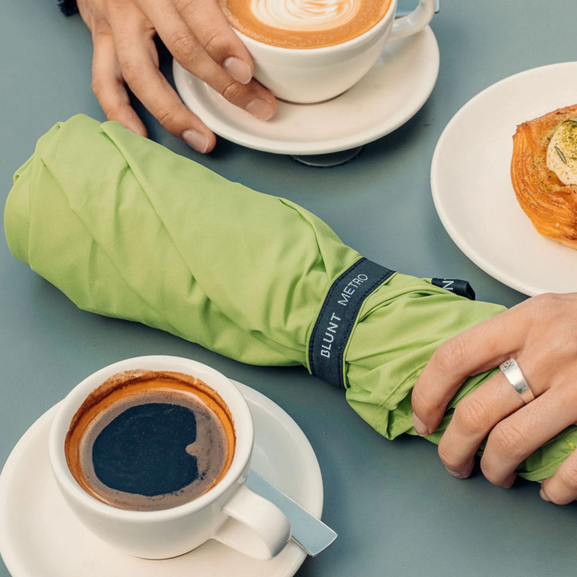 Retracted Blunt Umbrella Metro Meadow Green on a table which is compact, collapsible and convenient. Consist of the detachable UV fabric canopy, sturdy frame, shaft and handle. Fits in your handbag, tote or backpack. Built for fast living, those who travel light, and any weather.