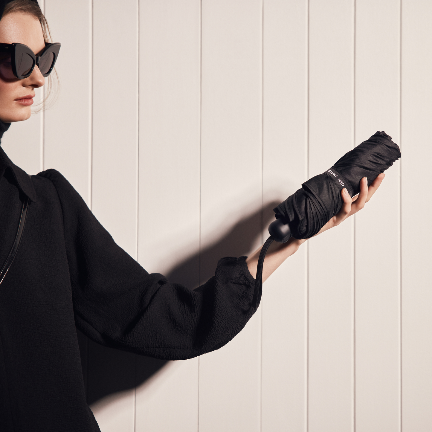 Folded Blunt Umbrella Black held by a woman, featuring a compact design with high tensioned canopy, wind tested construction and recycled rPET fabric for rain or shine.
