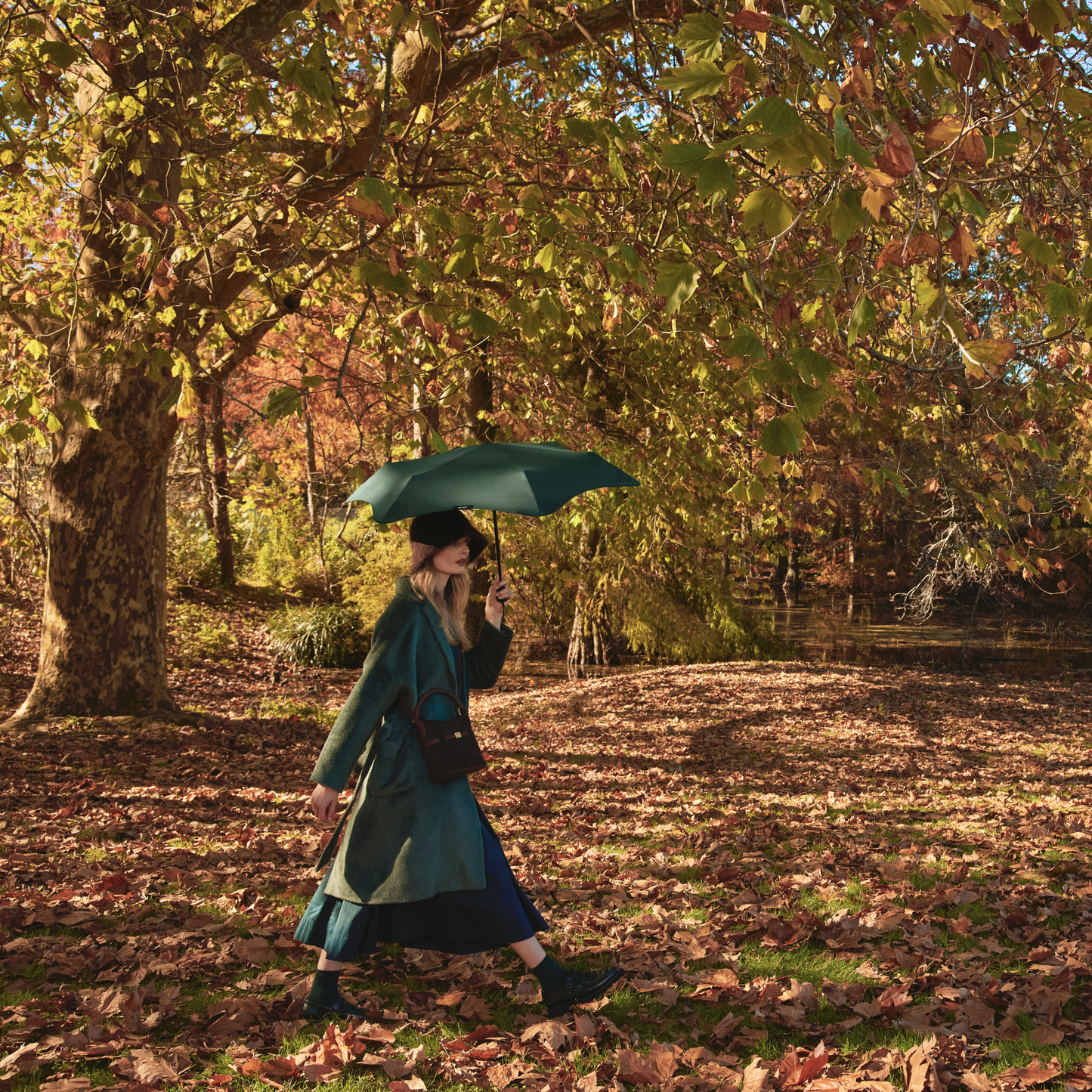 Side of the the woman in the forest holding the Blunt Umbrella Forest Green at 98cm, featuring a compact design with high tensioned canopy, wind tested construction and recycled rPET fabric for rain or shine.