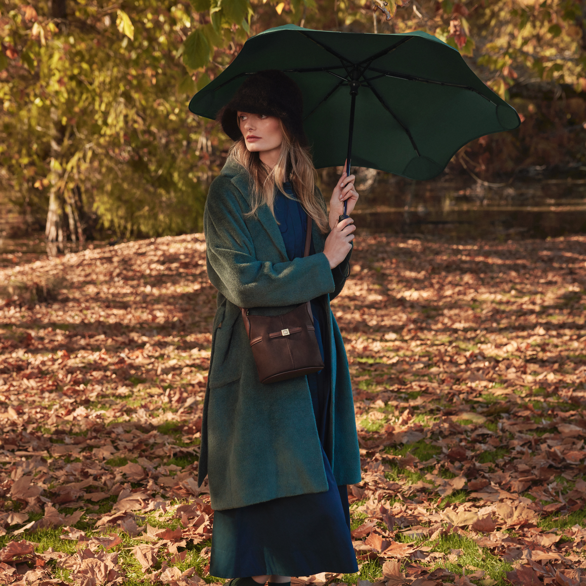 Front view of a woman holding the open Blunt Umbrella Forest Green at 98cm, featuring a compact design with high tensioned canopy, wind tested construction and recycled rPET fabric for rain or shine.