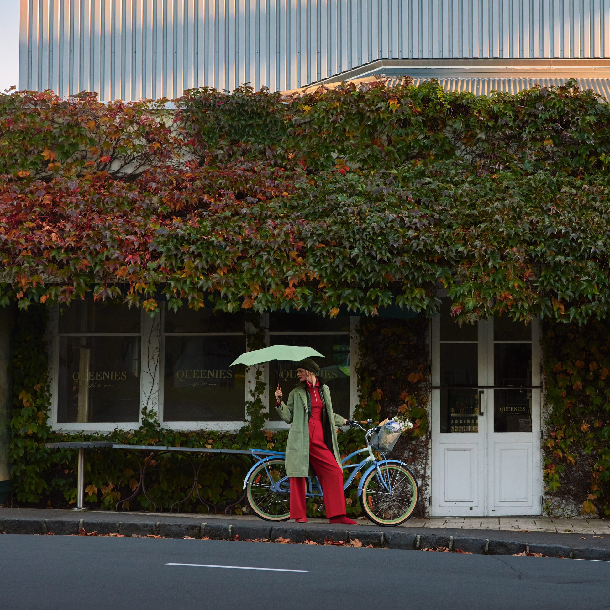 A woman parking her bicycle holding the open Blunt Umbrella Sage Green at 98cm, featuring a compact design with high tensioned canopy, wind tested construction and recycled rPET fabric for rain or shine.