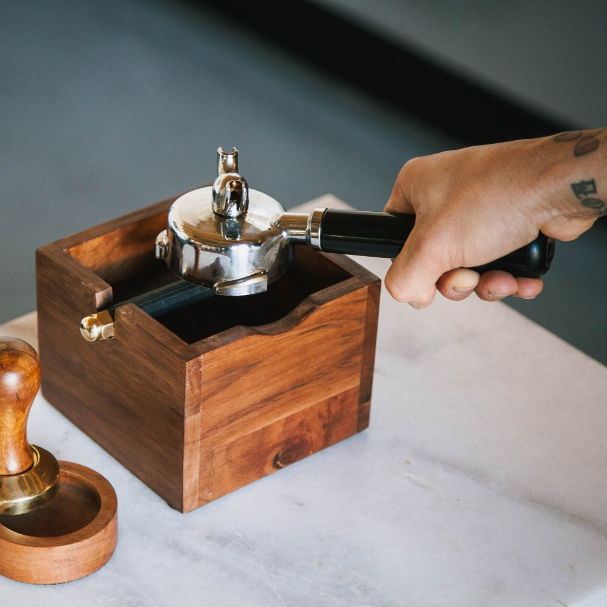 A hand taps a metal portafilter against the Brut Coffee Knock Box on a countertop, with a wooden coffee tamper nearby.