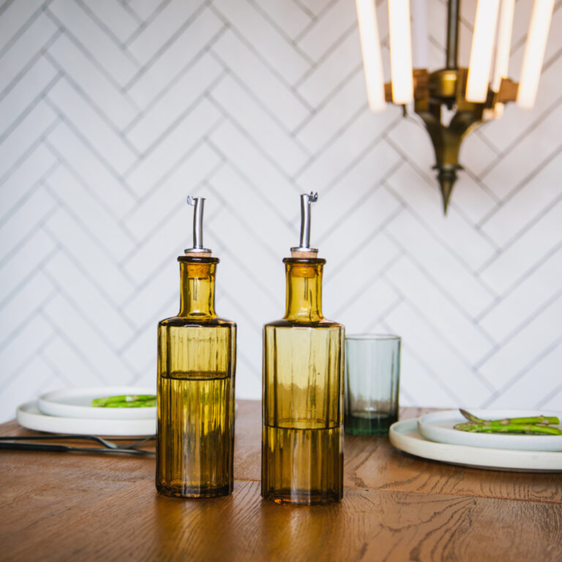 Shown on the table Brût Homeware Art Deco-inspired oil or vinegar bottle, amber, made from mouth-blown recycled glass with a natural cork pourer and anti-drip cap, handcrafted in India for a sustainable and elegant kitchen addition.