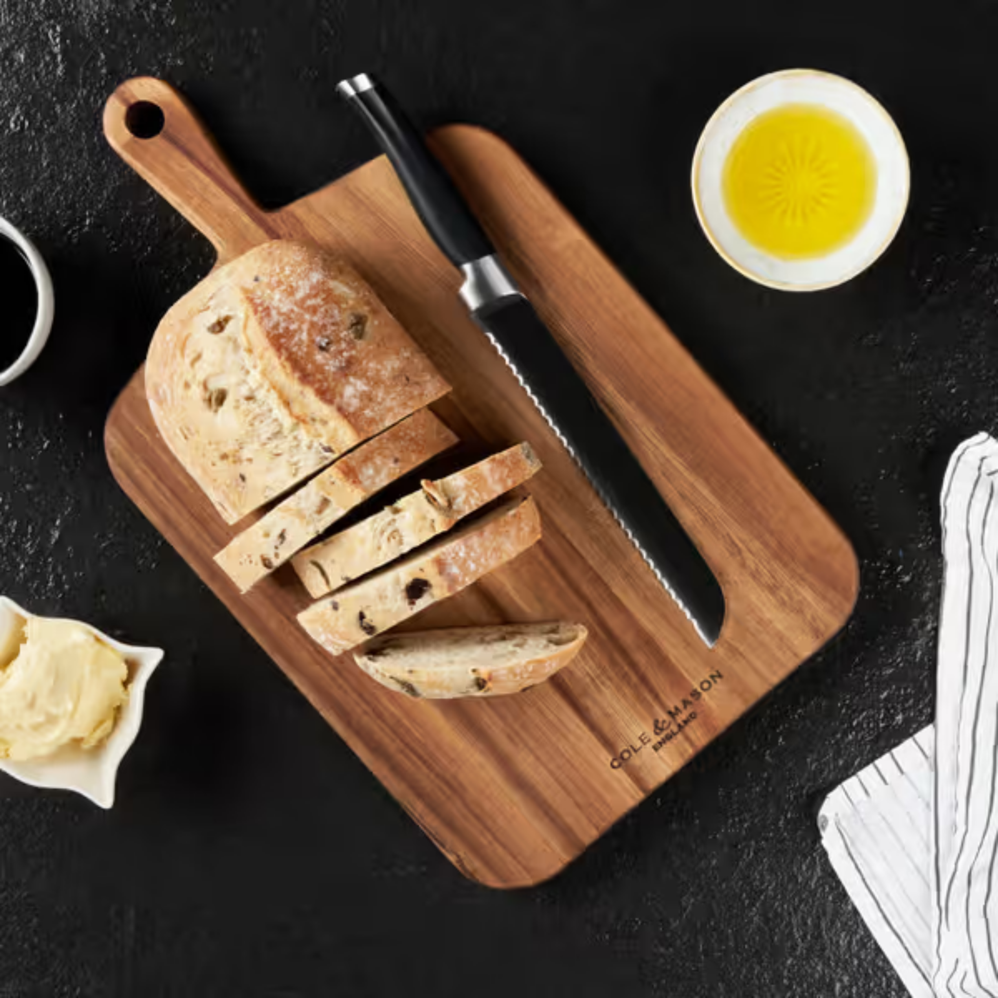Showing sliced bread with knife view of Cole & Mason medium double-sided acacia wood chopping board, natural finish, 2.5 cm thick, with handle and hanging hole, for chopping, slicing, and serving.