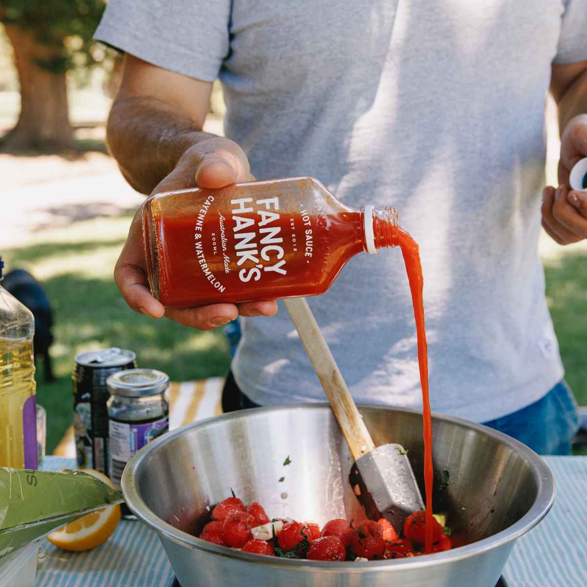 Pouring Fancy Hanks Cayenne & Watermelon Hot Sauce 200ml bottle, vibrant red sauce in a clear glass container with a white lid.