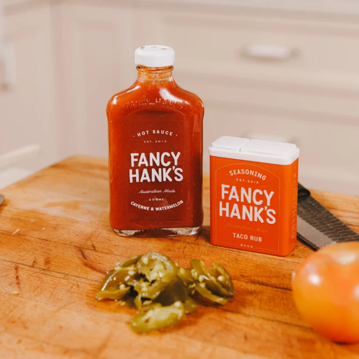 On the table, Fancy Hanks Cayenne & Watermelon Hot Sauce 200ml bottle, vibrant red sauce in a clear glass container with a white lid.