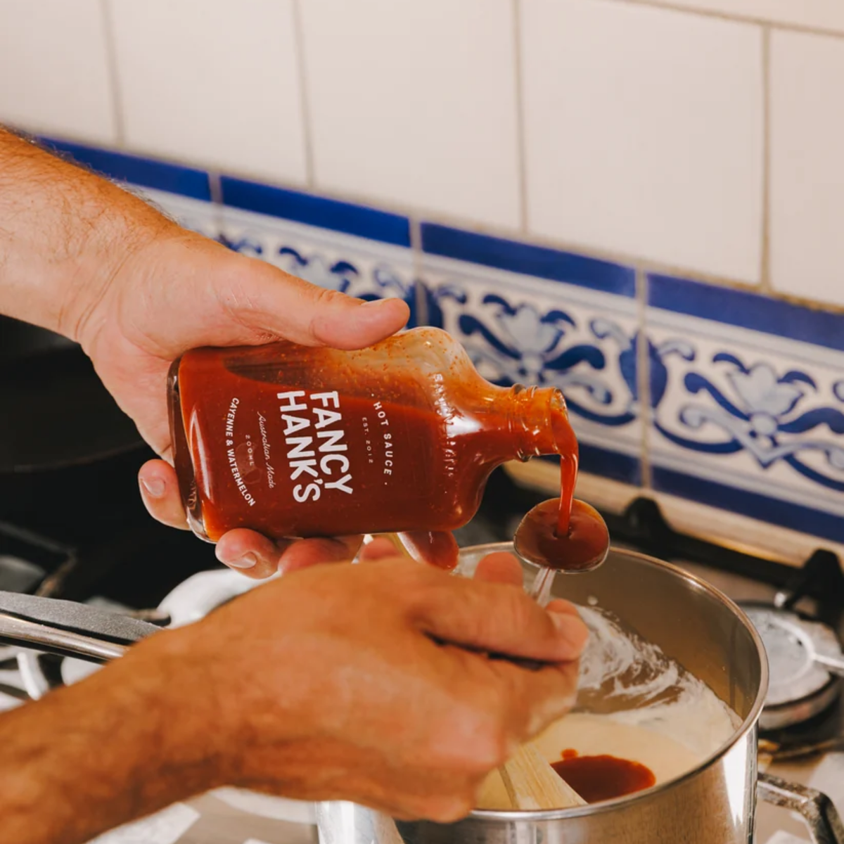 Putting Fancy Hanks Cayenne & Watermelon Hot Sauce to the pan from a 200ml bottle, vibrant red sauce in a clear glass container with a white lid.