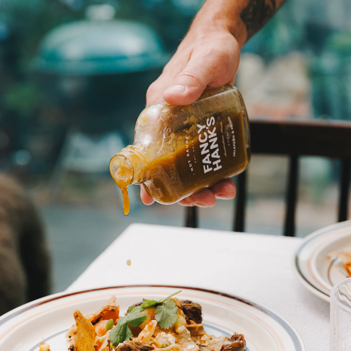 Pouring Fancy Hanks Jalapeno & Peach Hot Sauce on food from 200ml bottle, orange-yellow sauce in clear glass with white cap; sweet, tangy, medium heat, fruit-forward flavor.