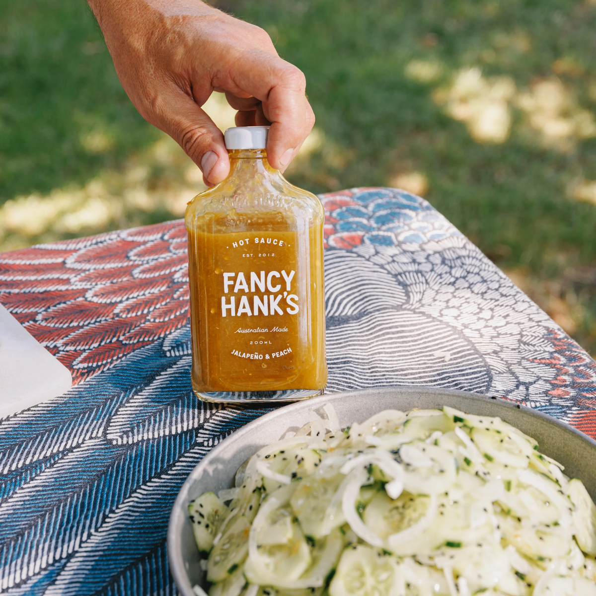 On the table, Fancy Hanks Jalapeno & Peach Hot Sauce 200ml bottle, orange-yellow sauce in clear glass with white cap; sweet, tangy, medium heat, fruit-forward flavor.