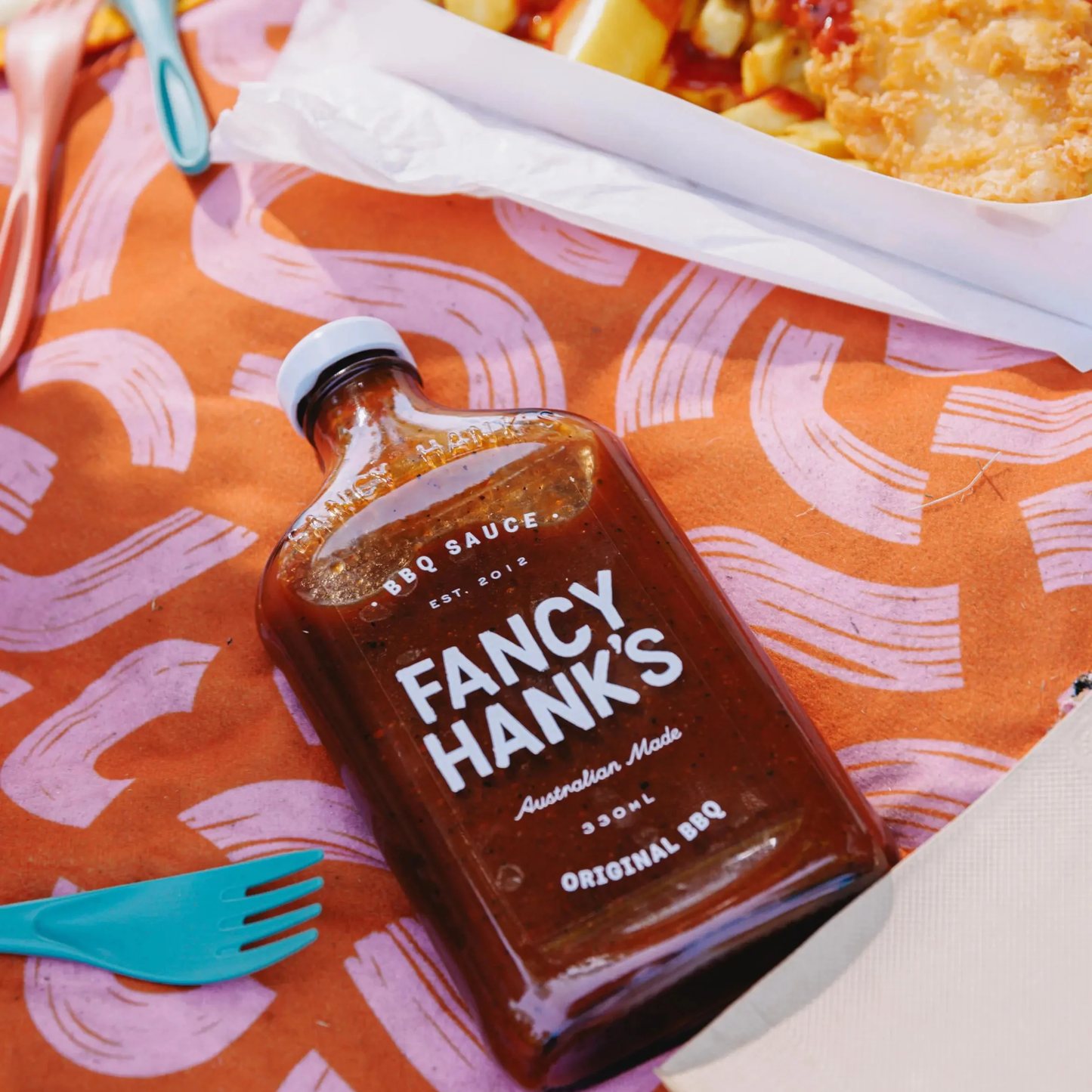Shown on a picnic mat, the Fancy Hanks Original BBQ Sauce 330ml, which is a slow-cooked American style barbecue sauce that is tangy, rich and molasses layered depth.