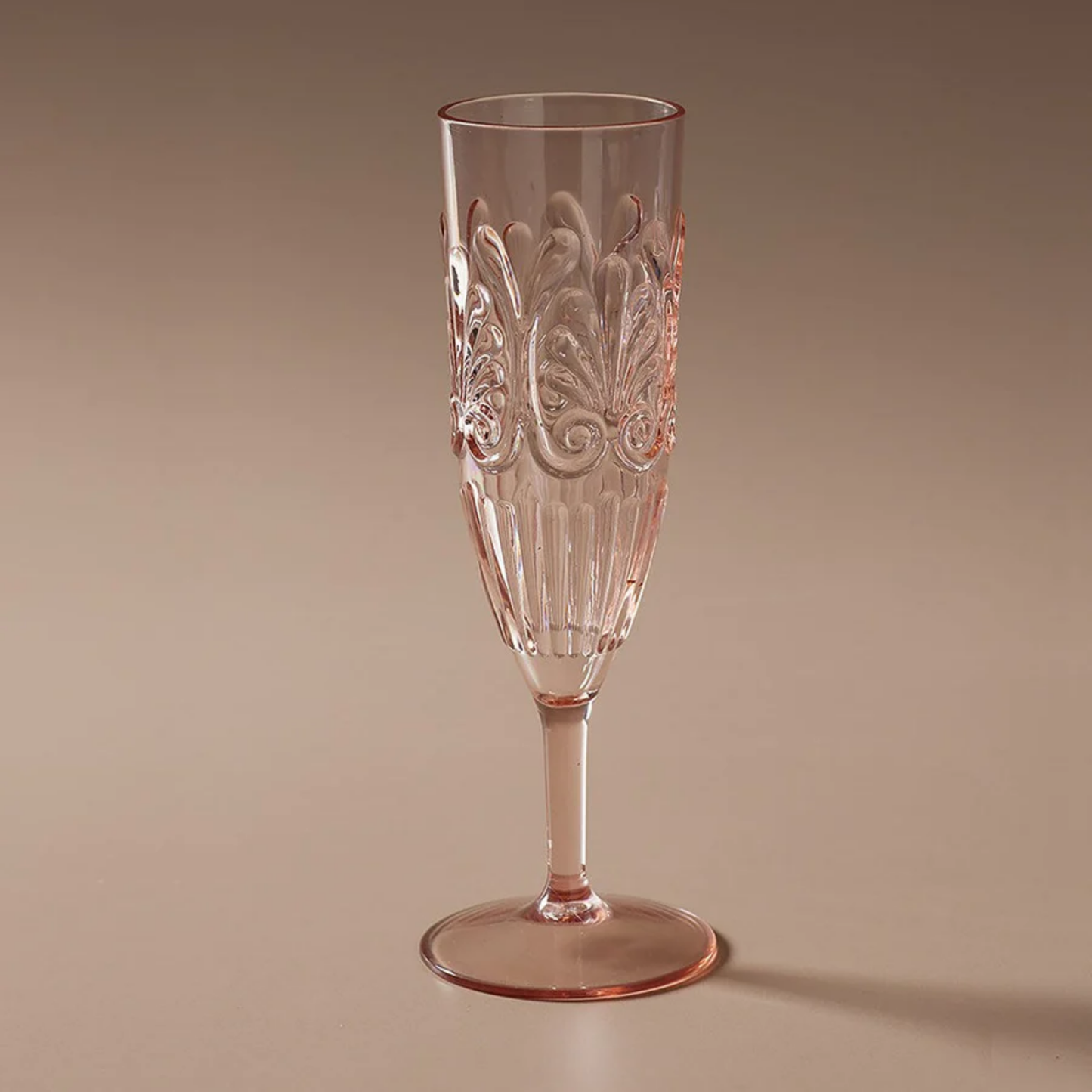 In a brown background Pale pink Fathom Clear acrylic champagne flute with elegant embossed detailing, glass-like clarity, and shatter-resistant BPA-free material.