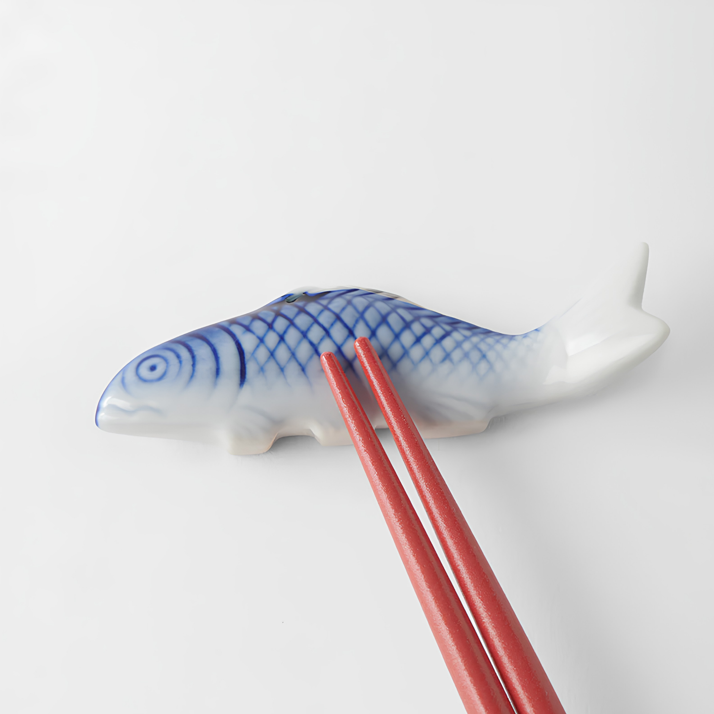 Laid on a surface with a pair of red chopsticks on top of the Fathom Chopstick Rest Blue Fish that has slight irregularities in size, glaze and texture from the artisan of the product
