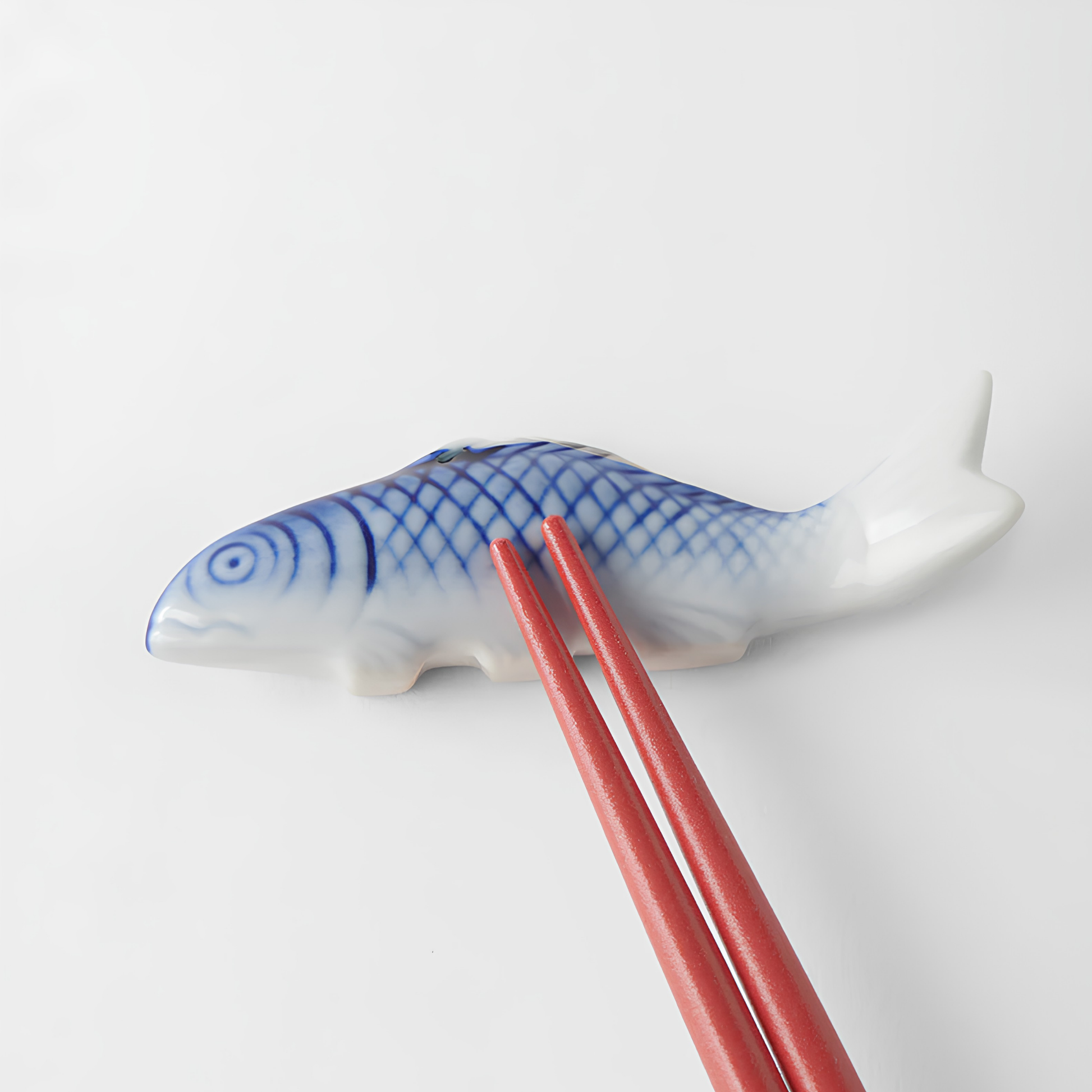 Laid on a surface with a pair of red chopsticks on top of the Fathom Chopstick Rest Blue Fish that has slight irregularities in size, glaze and texture from the artisan of the product