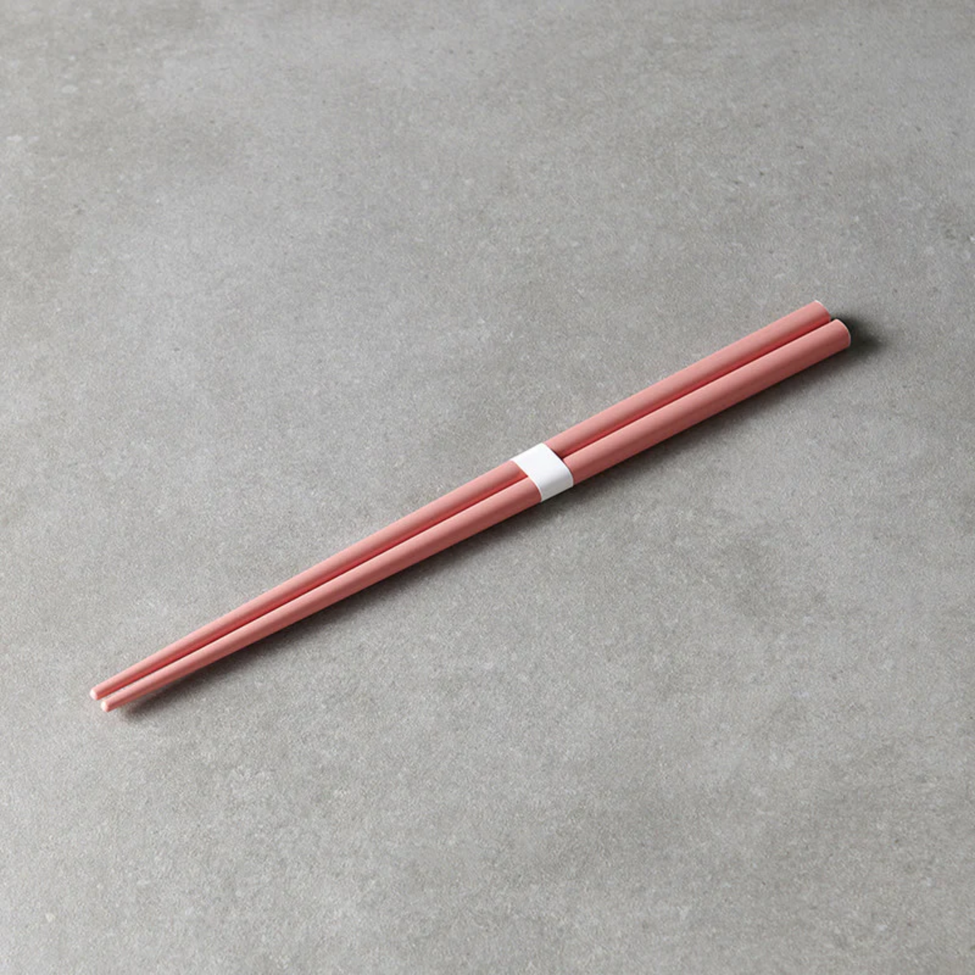Laid on a gray surface the Fathom Chopsticks Pair Lacquered Pink, soft finish with sleek white accent.