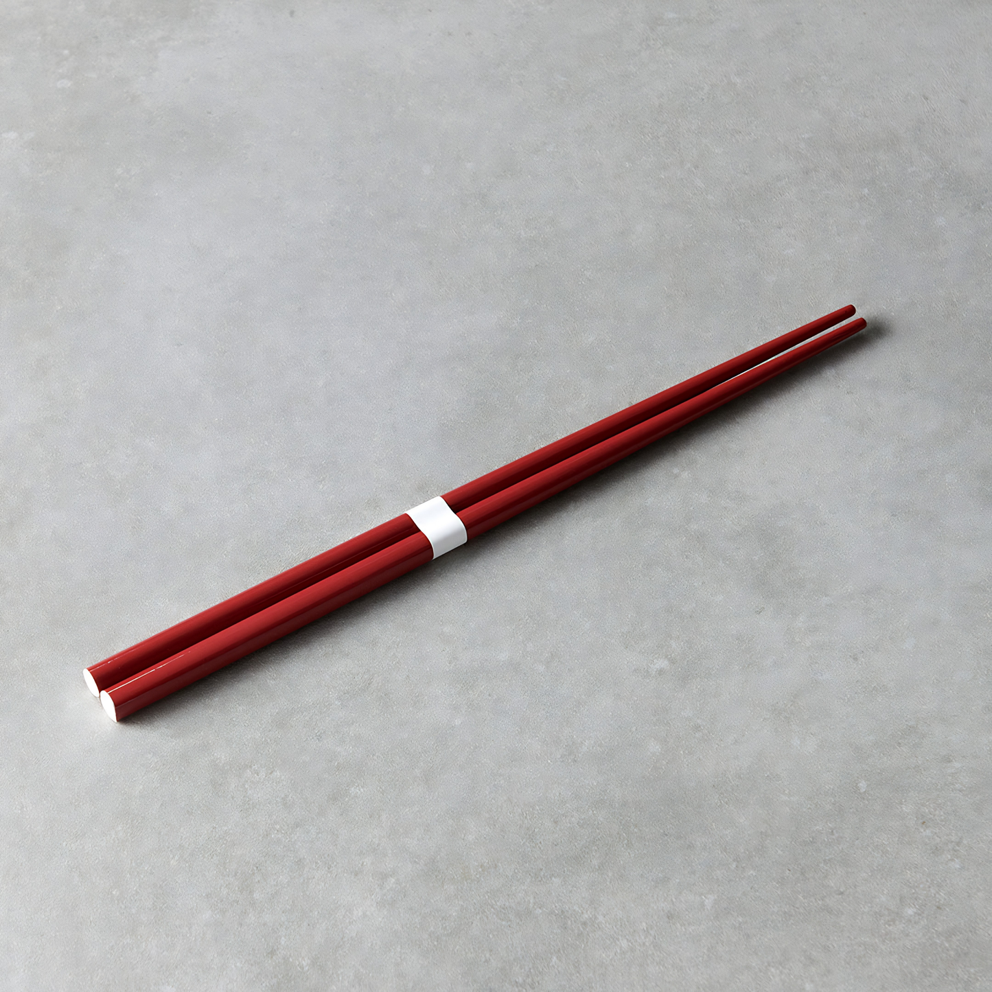 Laid on a gray surface the Fathom Chopsticks Pair Lacquered Red deep finish with white accent.