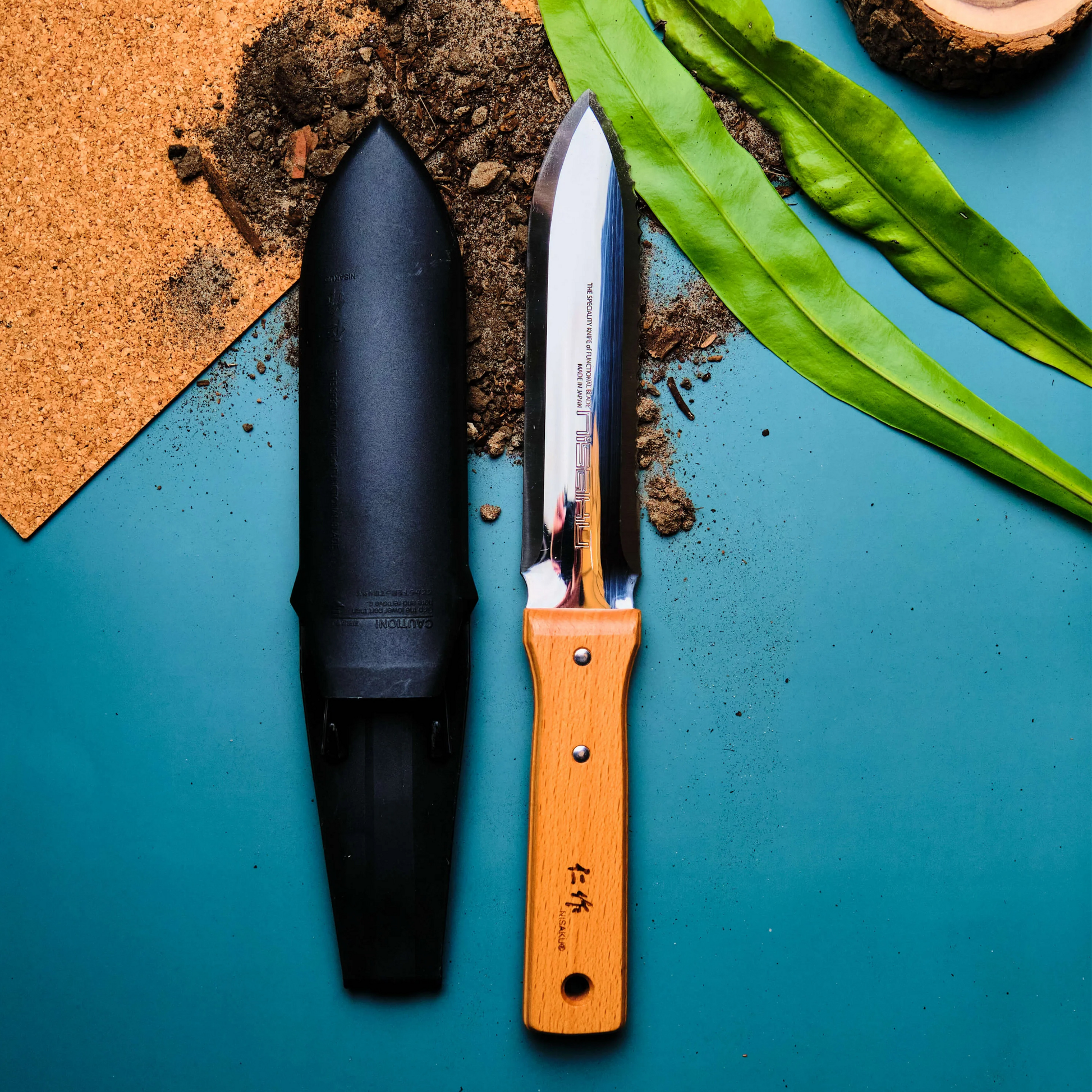 Fathom Hori Hori Japanese Garden Knife with Sheath on a blue surface with soul. Features a dual edged that is one serrated and one straight with scalloped stainless steel, hardwood handle and hard sheath.