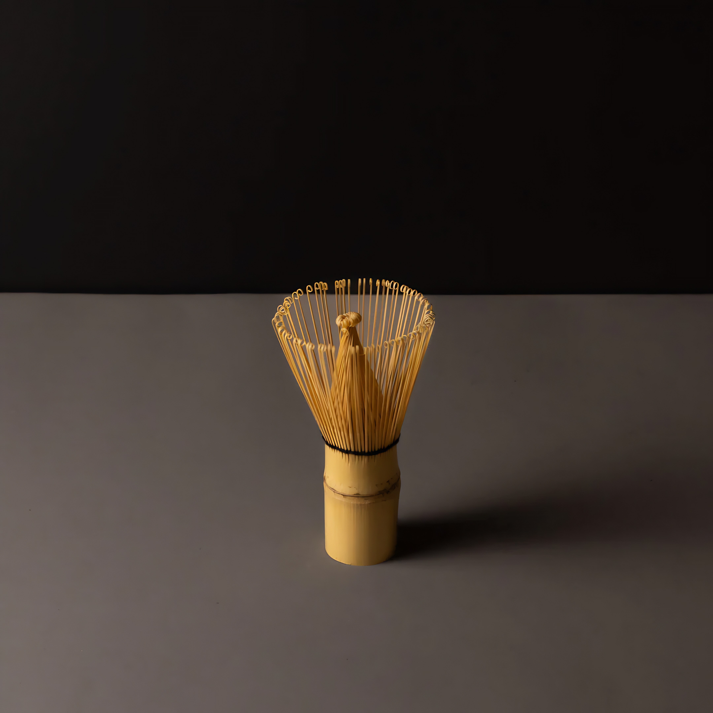 Standing on a surface with a black background the Fathom Matcha Whisk Bamboo 100 Prong, Chasen handcrafted from a single bamboo into delicate bristles making it as whisk