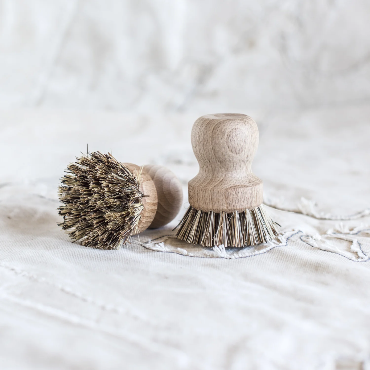 Fathom Pot Brush Tampico and Palmyra Fibre- Raw beechwood brushes with stiff bristles rest on textured fabric.