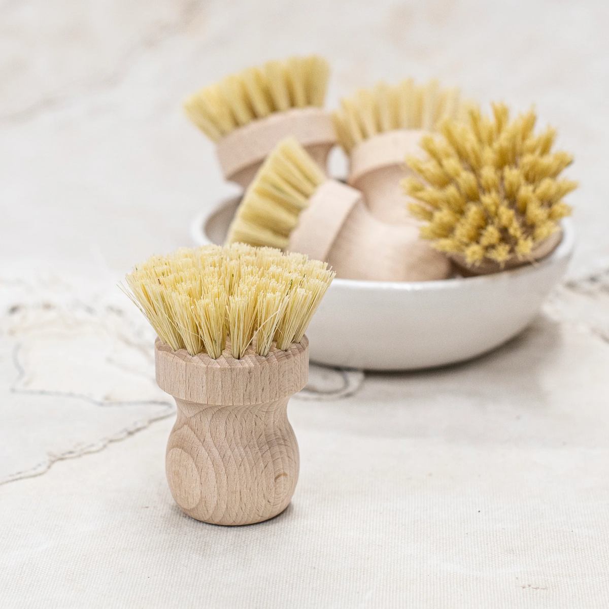 Fathom Pot Brush Tampico Fibre- Raw beechwood brushes with stiff bristles rest on textured fabric.