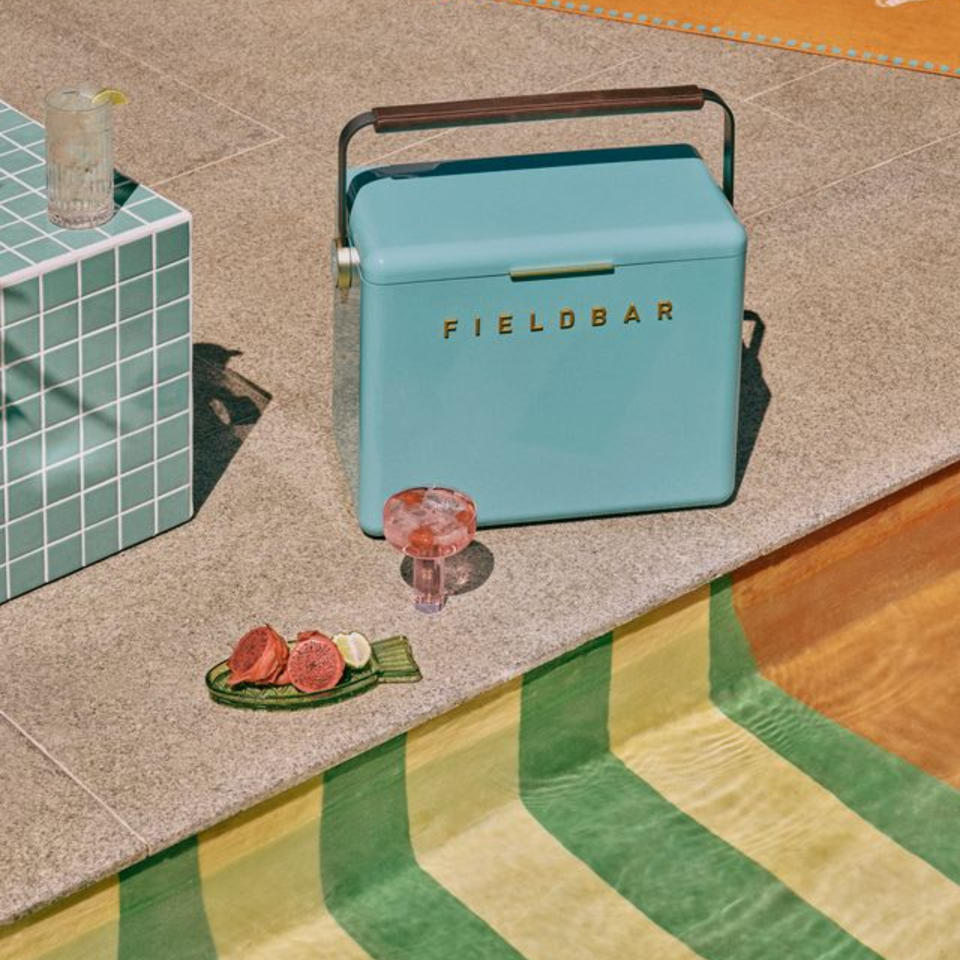 FIELDBAR Gin Trunk Bazaruto Blue shown on the poolside with drinks and fruits- premium hardcase 24L cooler box in impact-resistant material with leather-wrapped handle, extruded aluminum carry handle, reflective heat-reducing finish, airtight gasket, BPA-free insulated interior, and 50-hour ice performance.