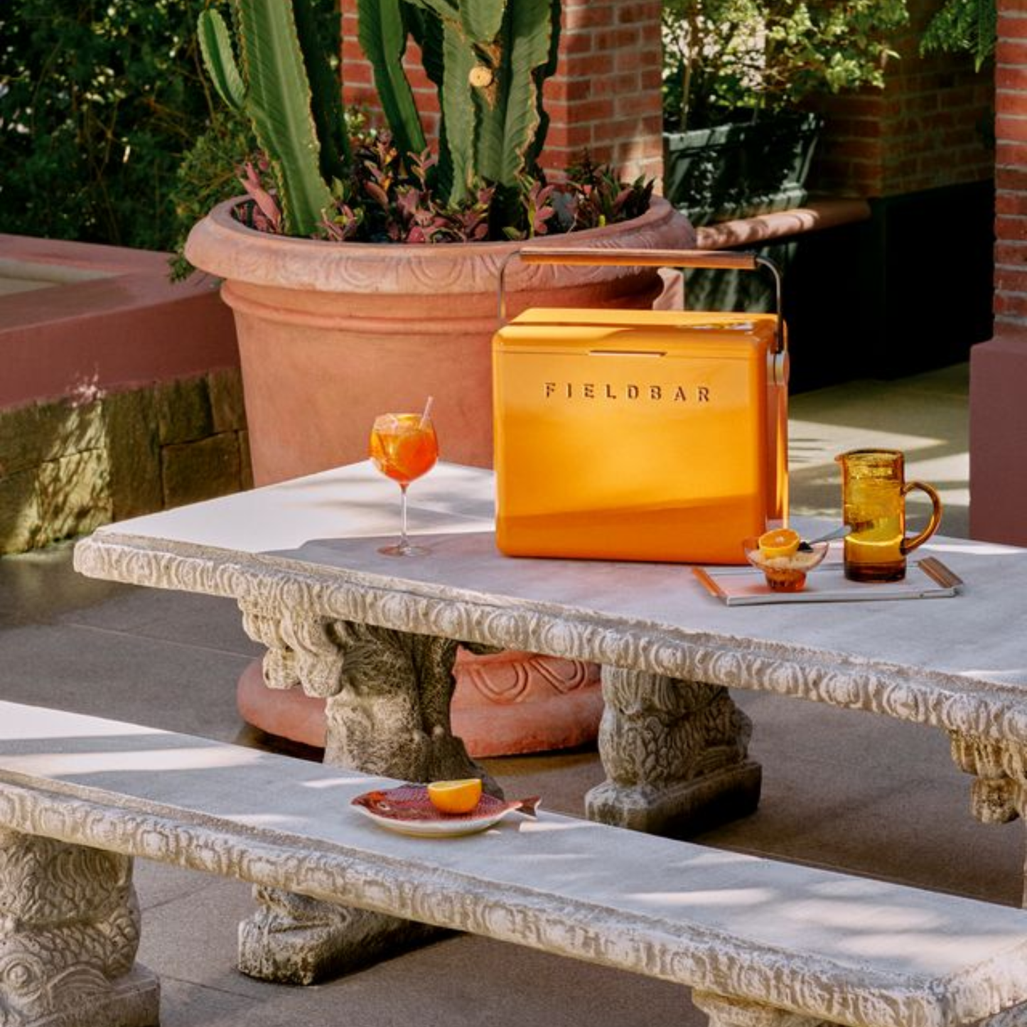 FIELDBAR Gin Trunk Orchard Orange shown on the table with drinks and food - premium hardcase 24L insulated cooler box in impact-resistant material with leather-wrapped handle, extruded aluminum carry handle, reflective heat-reducing finish, airtight water- and air-sealed gasket, BPA-free interior, removable foam insulation, and 50-hour ice performance.