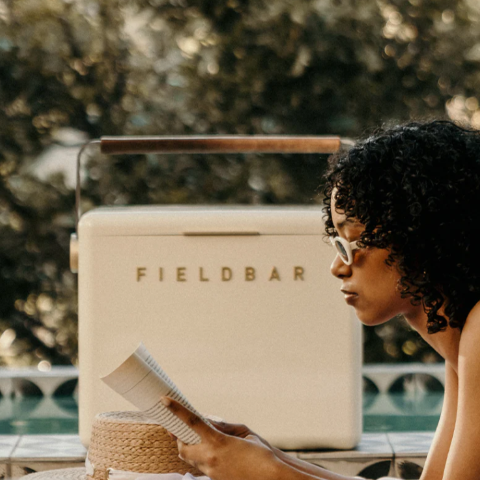 Front view of FIELDBAR Gin Trunk Safari White on a poolside with a woman reading a book, showing size comparison to the girl - premium hardcase 24L insulated cooler box in white impact-resistant material with leather-wrapped handle, extruded aluminum carry handle, reflective heat-reducing finish, airtight water- and air-sealed gasket, BPA-free interior, removable high-performance insulation, and 50-hour ice retention.