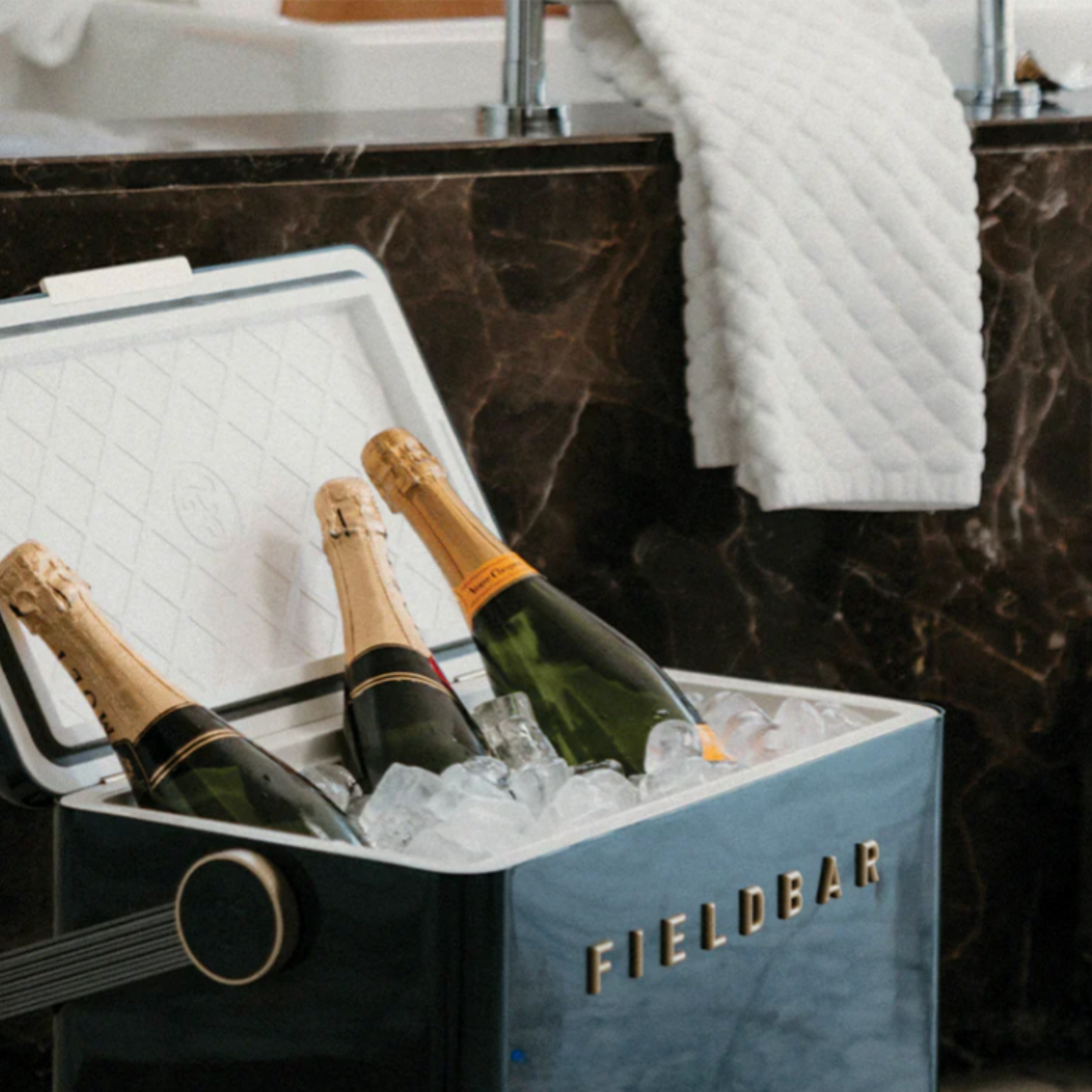 FIELDBAR Gin Trunk Sea Boat Blue shown with 3 bottles of wine inside with ice cubes- premium hardcase 24L insulated cooler box in blue impact-resistant material with leather-wrapped handle, extruded aluminum carry handle, reflective heat-reducing paint, airtight water- and air-sealed gasket, BPA-free easy-clean interior, removable high-performance insulation, and 50-hour ice retention.